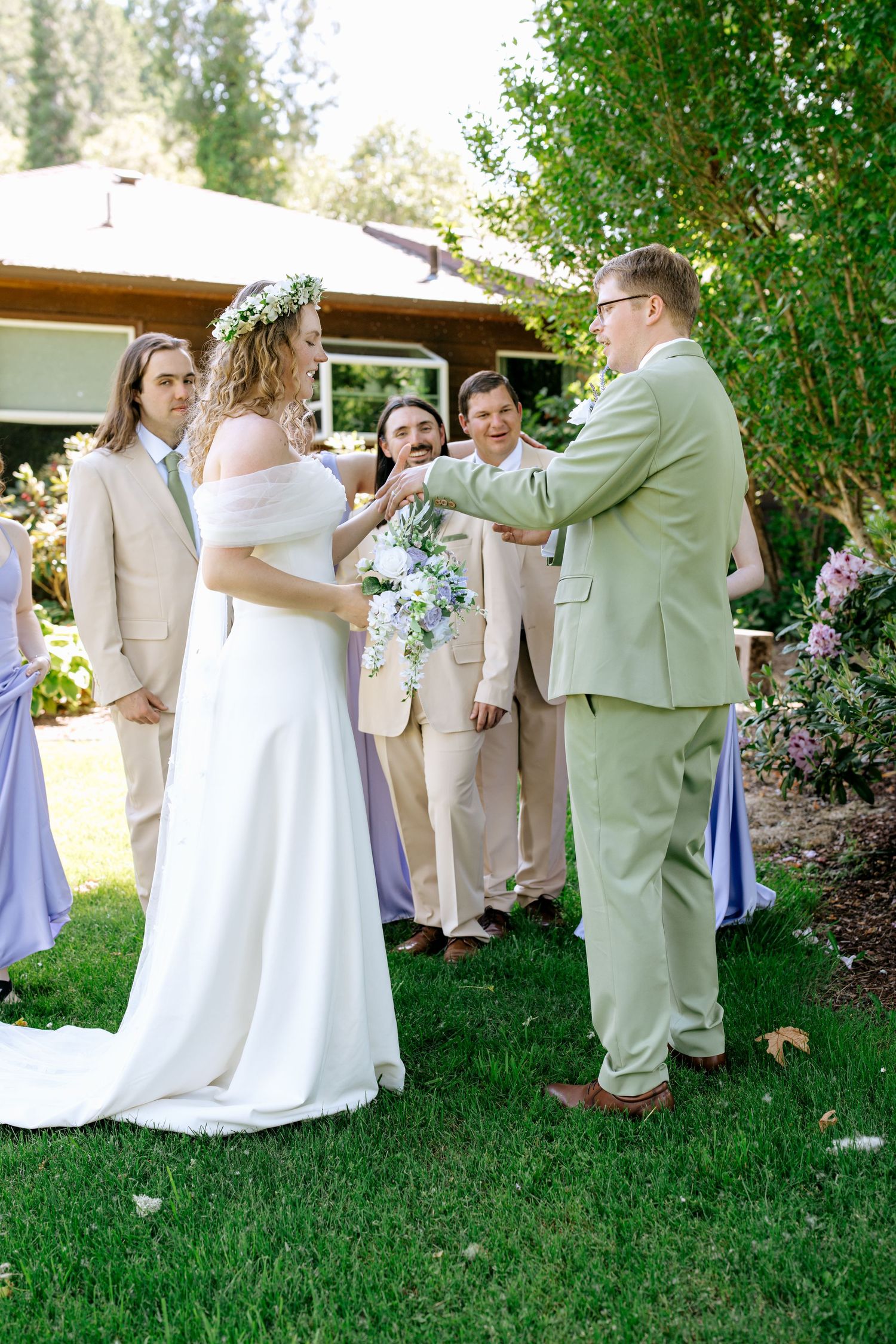 Bride in white gown with flower crown exchanges vows with groom in sage green suit surrounded by wedding party outdoors.