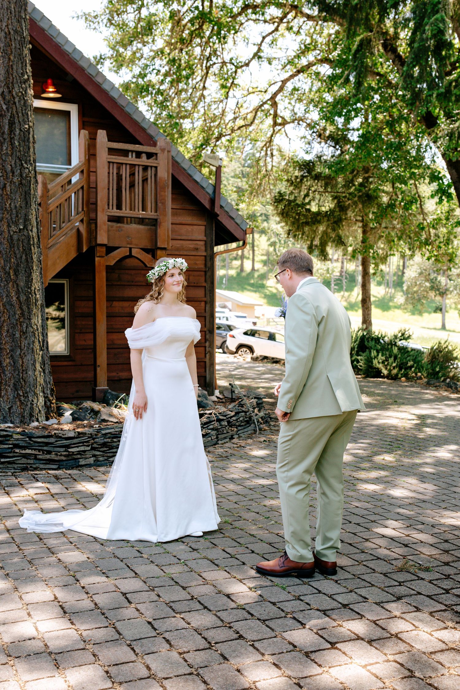Bride in white off-shoulder gown with floral crown meets groom in tan suit for first look outside rustic wooden cabin.