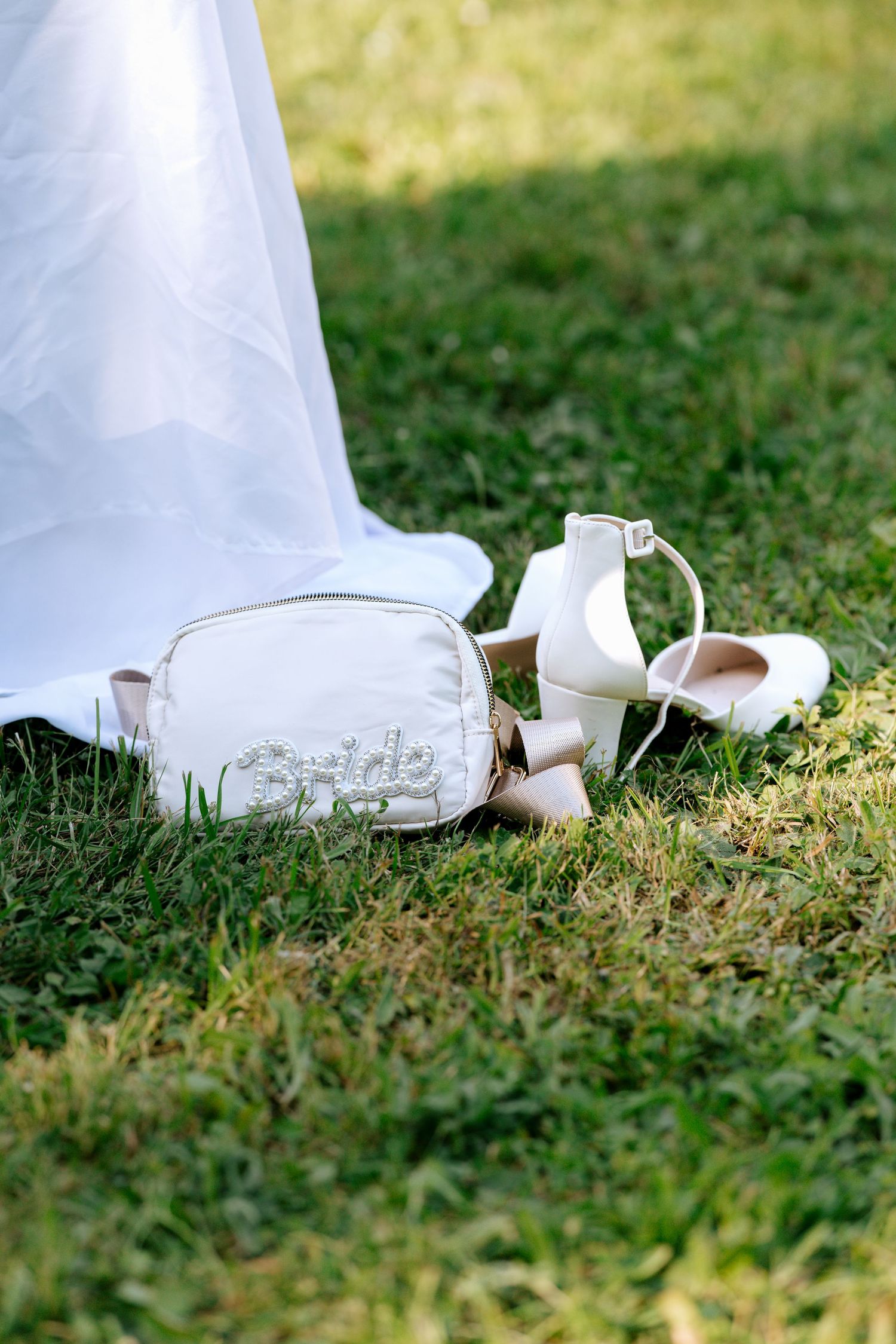 Bridal heels and bride bag accessories arranged on green grass beneath flowing white wedding gown.