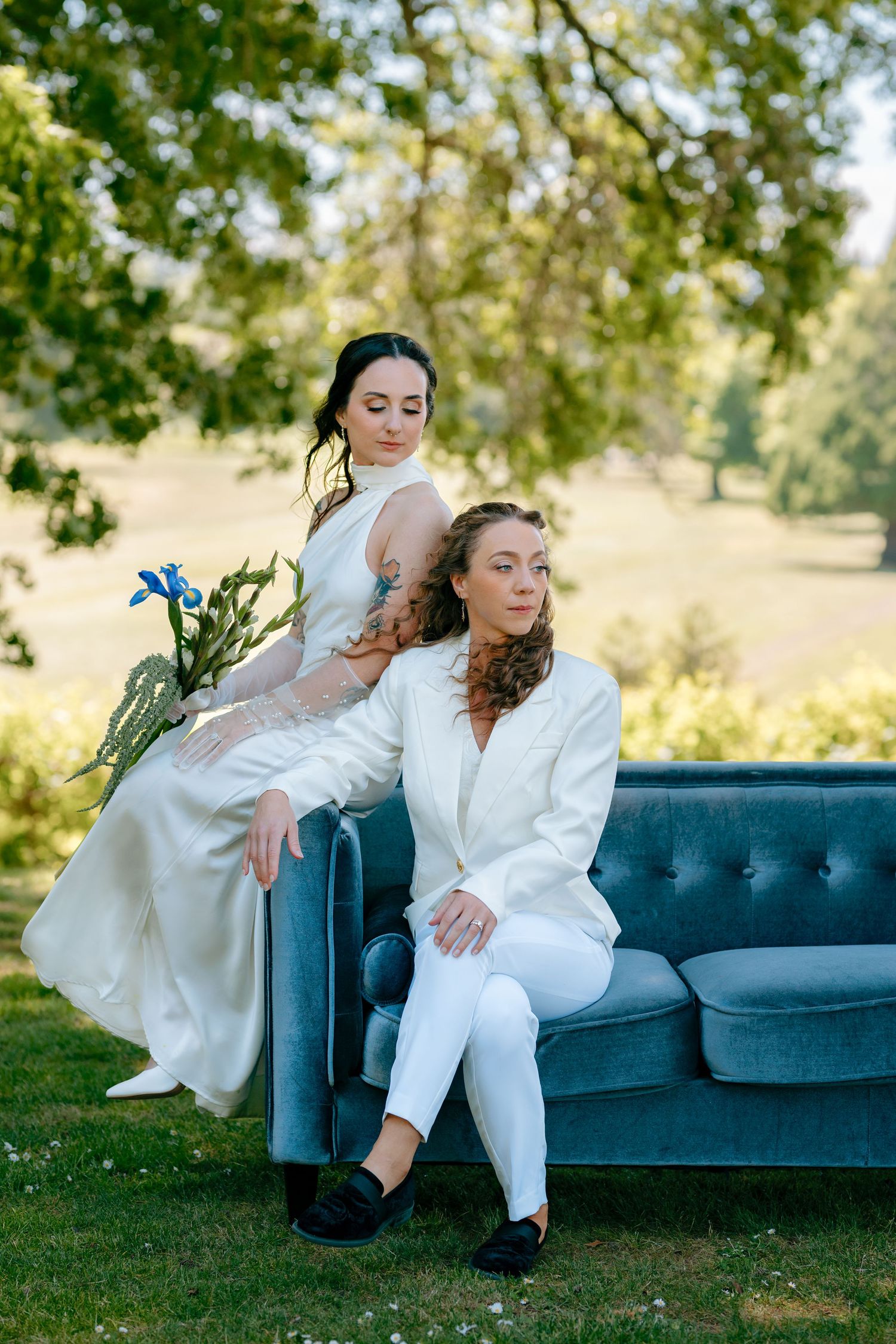 Two brides in white attire pose on a blue velvet sofa outdoors, surrounded by lush green trees.