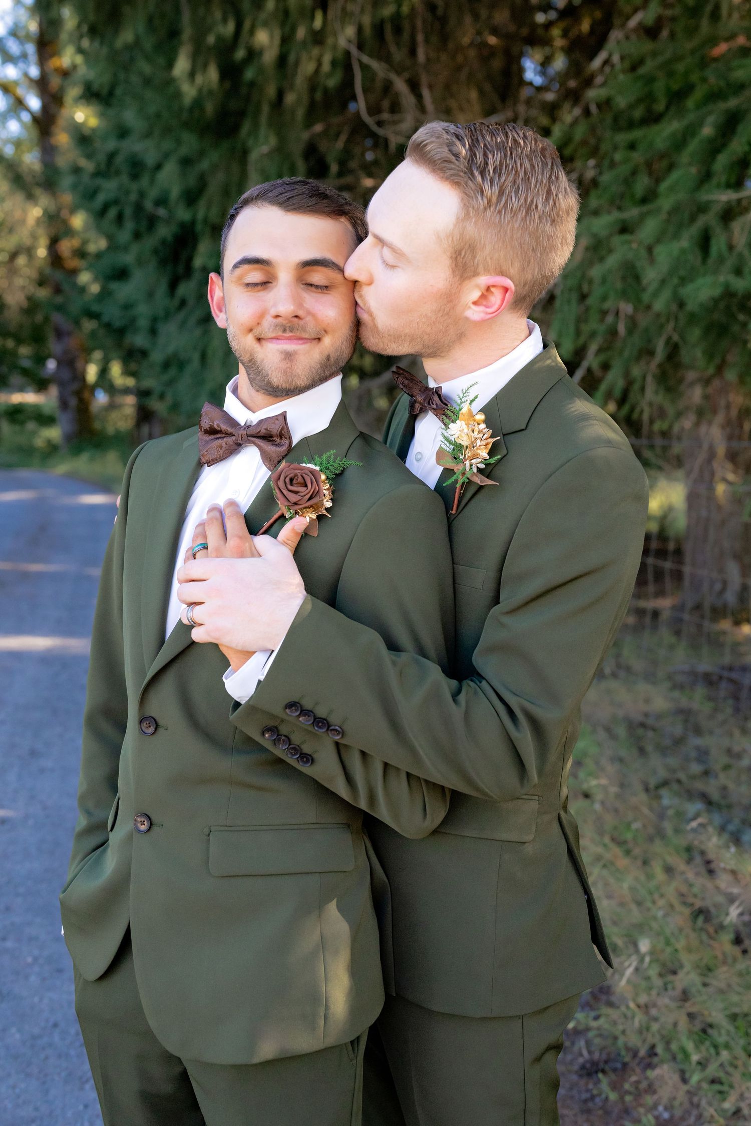 Two grooms in matching olive green suits share a tender kiss on the cheek during their outdoor wedding portrait session.
