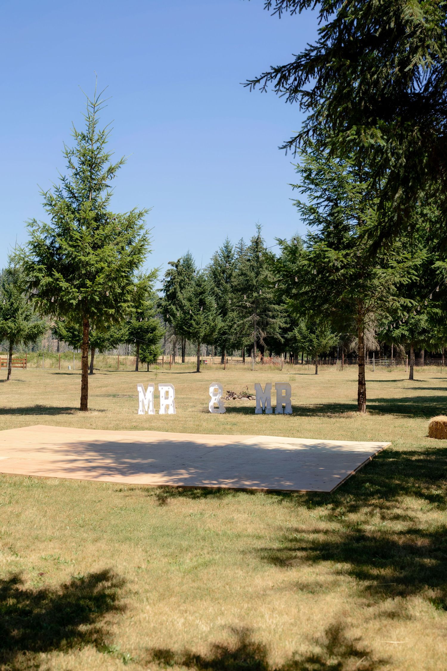 Outdoor wedding venue with Mr & Mr marquee letters on a dance floor surrounded by tall evergreen trees.