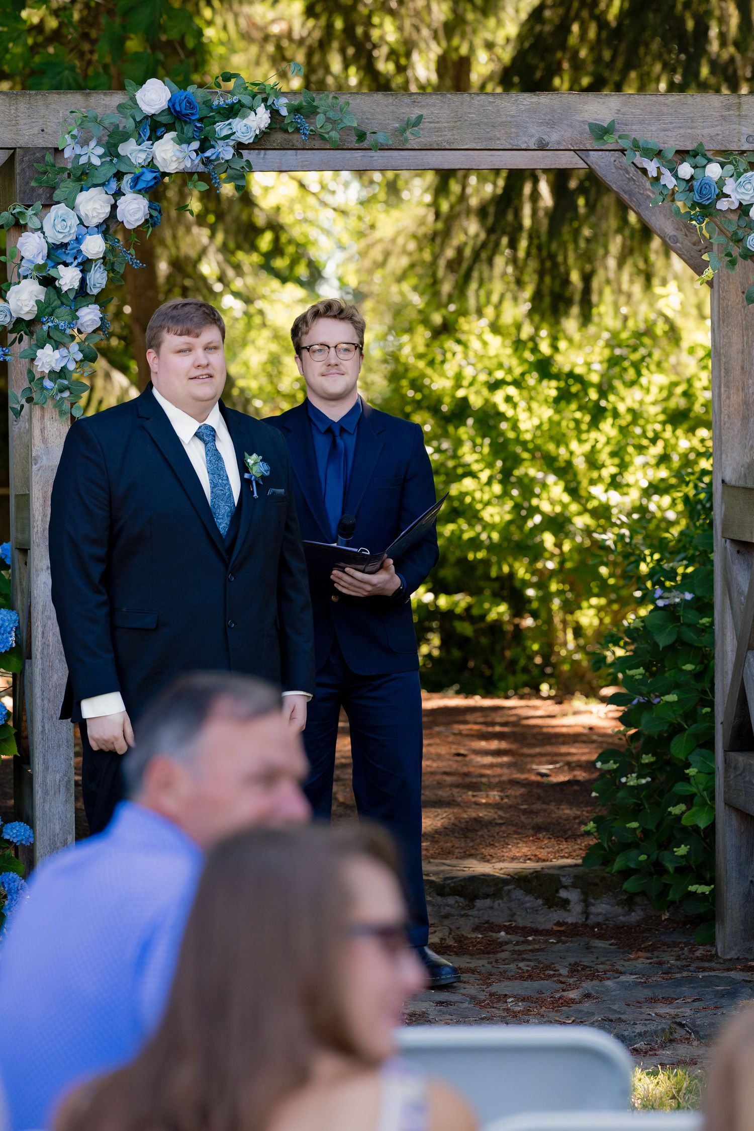 Groom and officiant stand under floral wooden arch awaiting bride at outdoor wedding ceremony with seated guests.