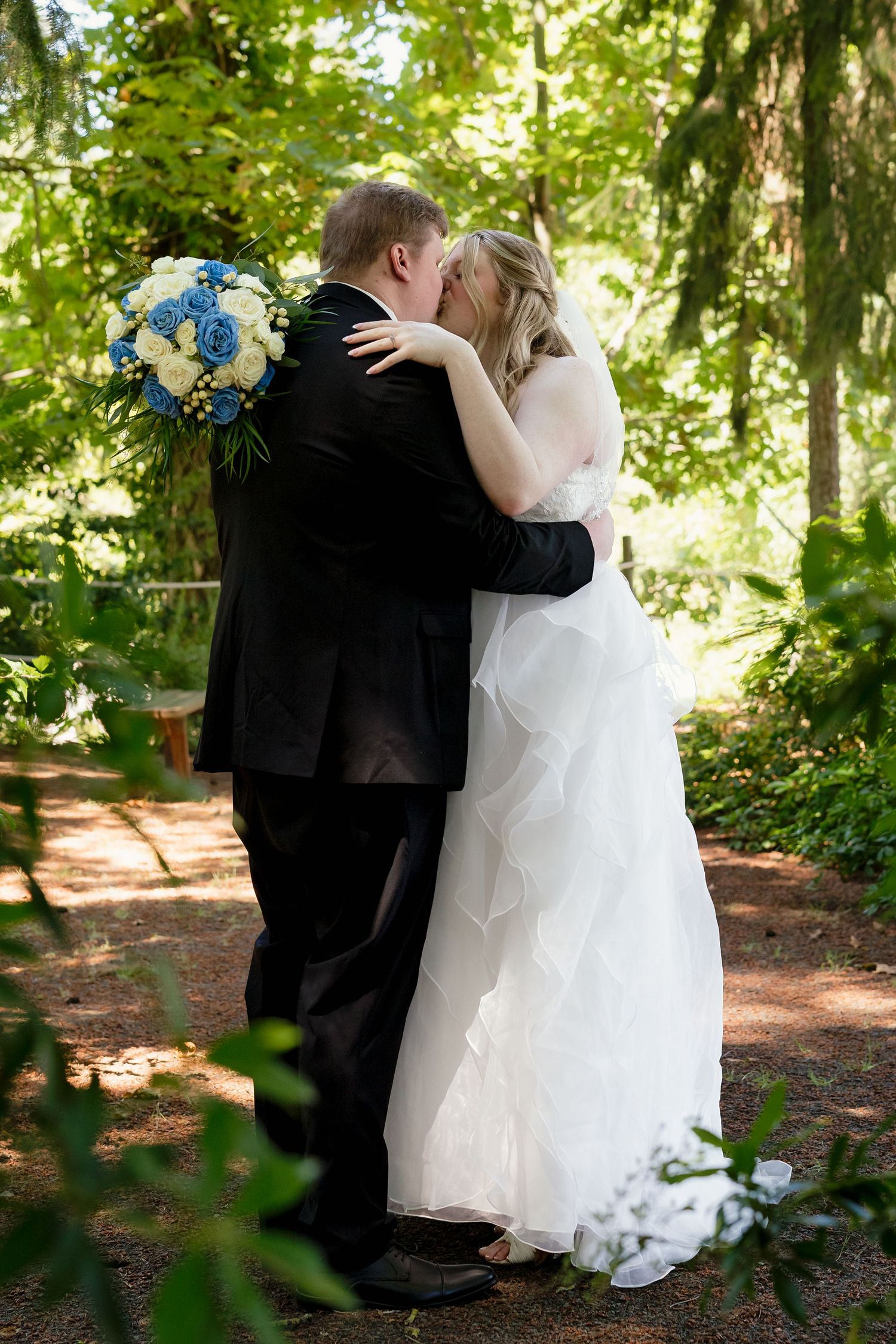 Bride and groom sharing their first kiss as a married couple in a lush green forest setting, bride holding blue and white bouquet.