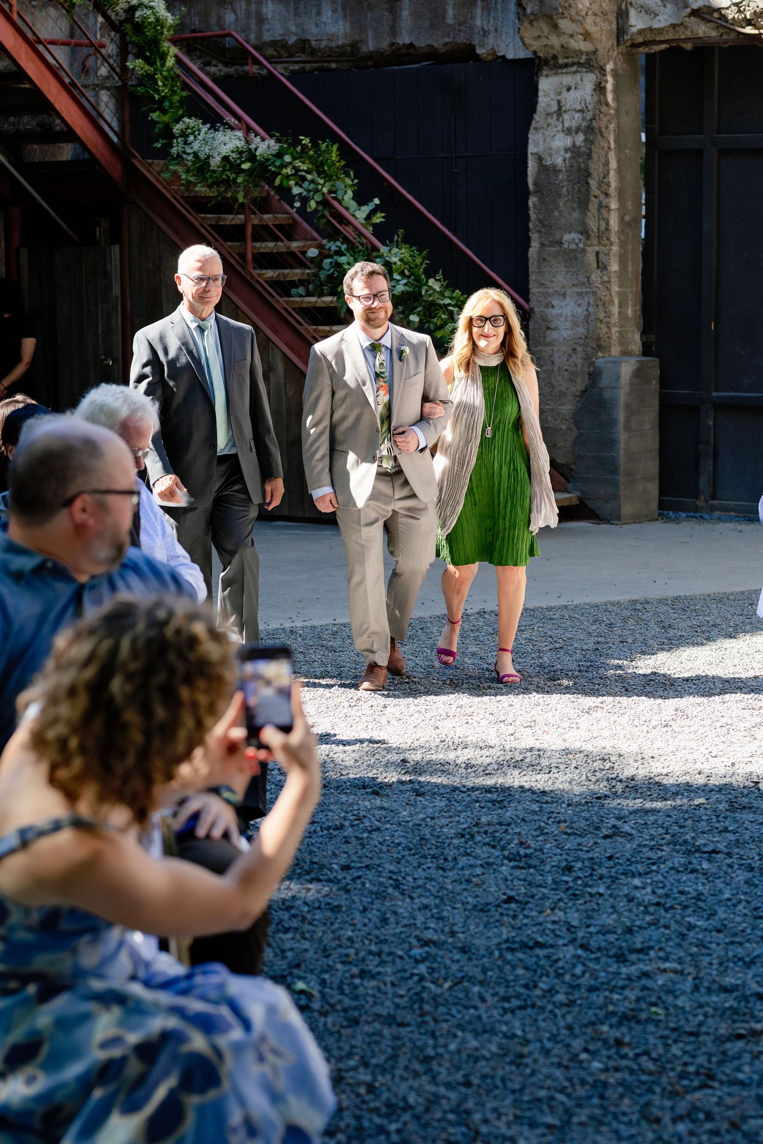 Wedding guests walk through industrial courtyard venue while attendee photographs the procession.