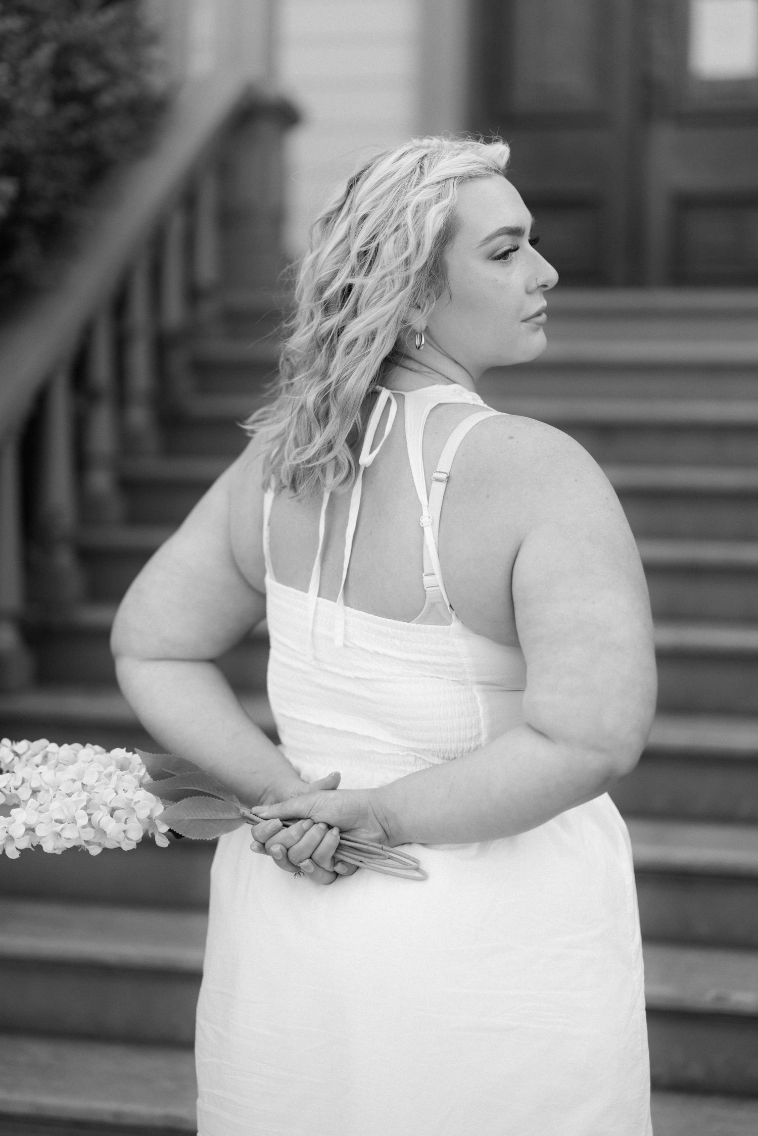 Black and white photo of a plus-size bride in white halter dress holding bouquet, posing on steps.