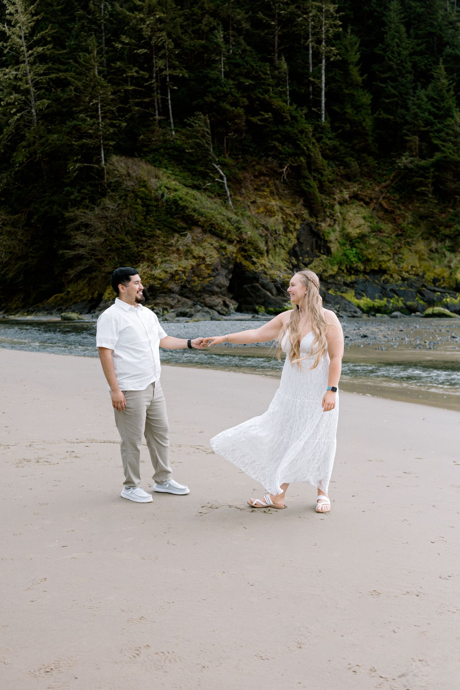 Couple holding hands and dancing on a sandy beach with lush green forest and rocky cliffs in the background.