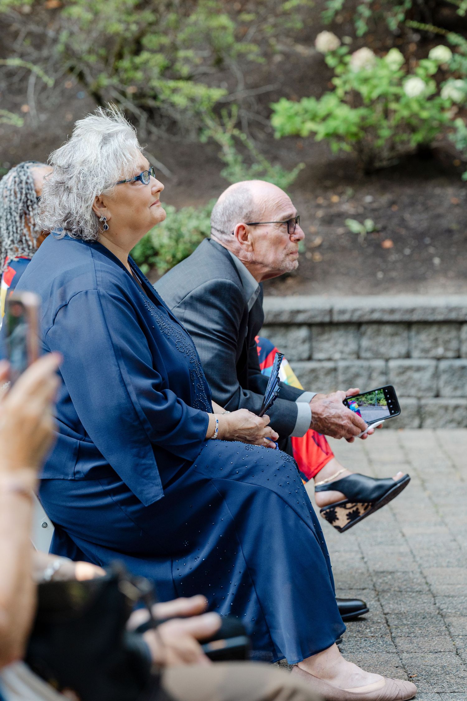 Elderly wedding guests seated outdoors, one holding a smartphone to capture the ceremony.