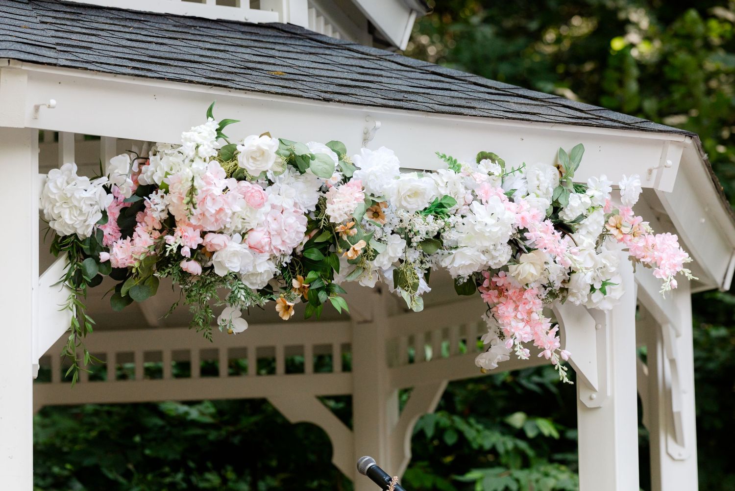 White gazebo adorned with lush pink and white floral arrangement featuring hydrangeas and greenery for a wedding ceremony.
