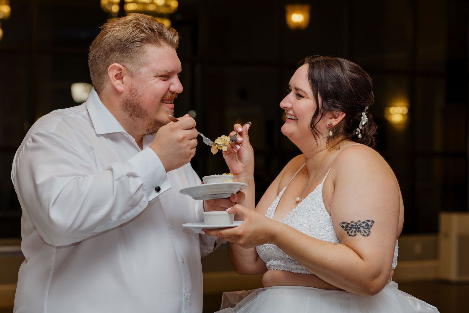 Smiling bride and groom share a bite of wedding cake together at their reception indoors.