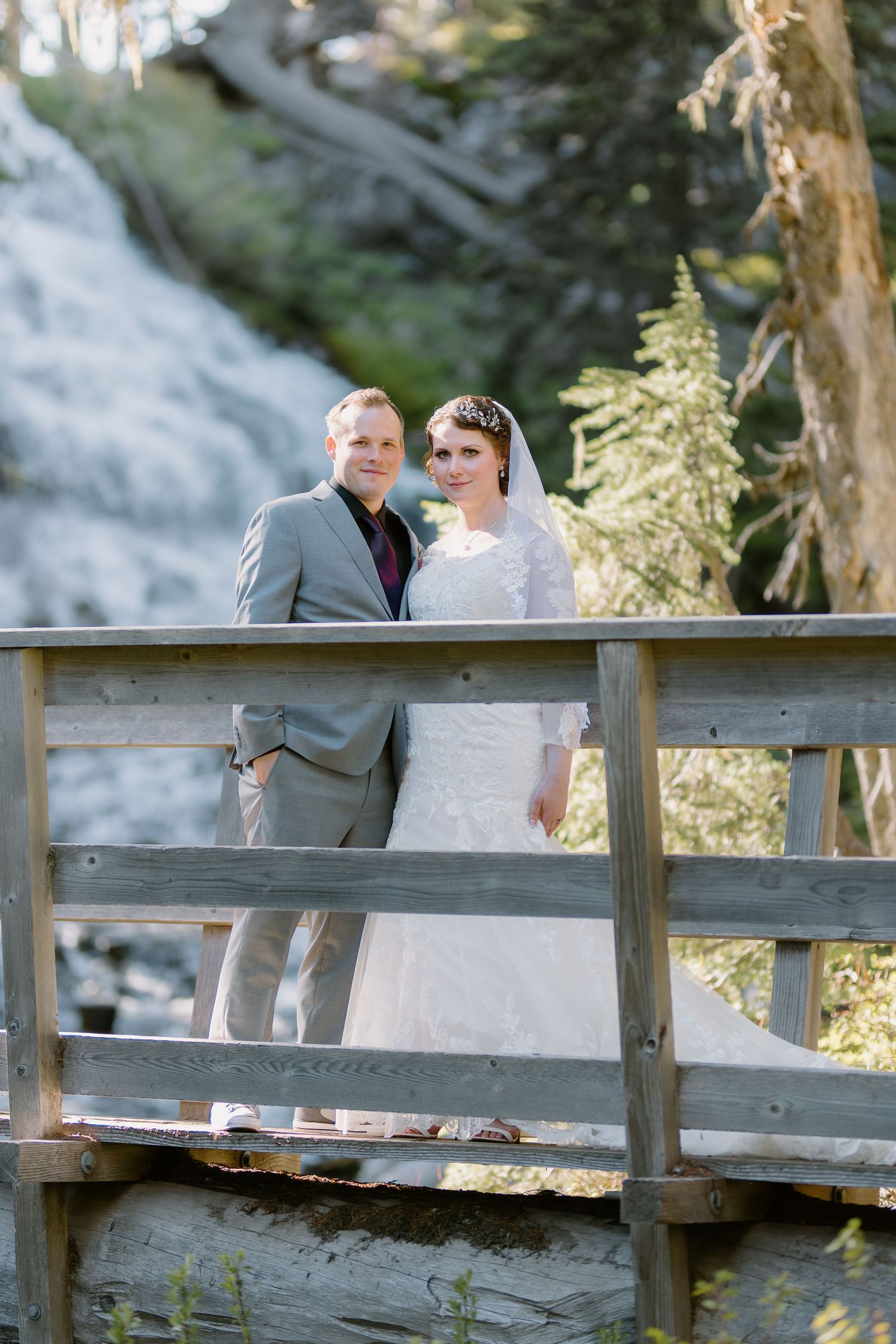 Bride and groom pose on a wooden bridge with a scenic waterfall and lush trees in the background.