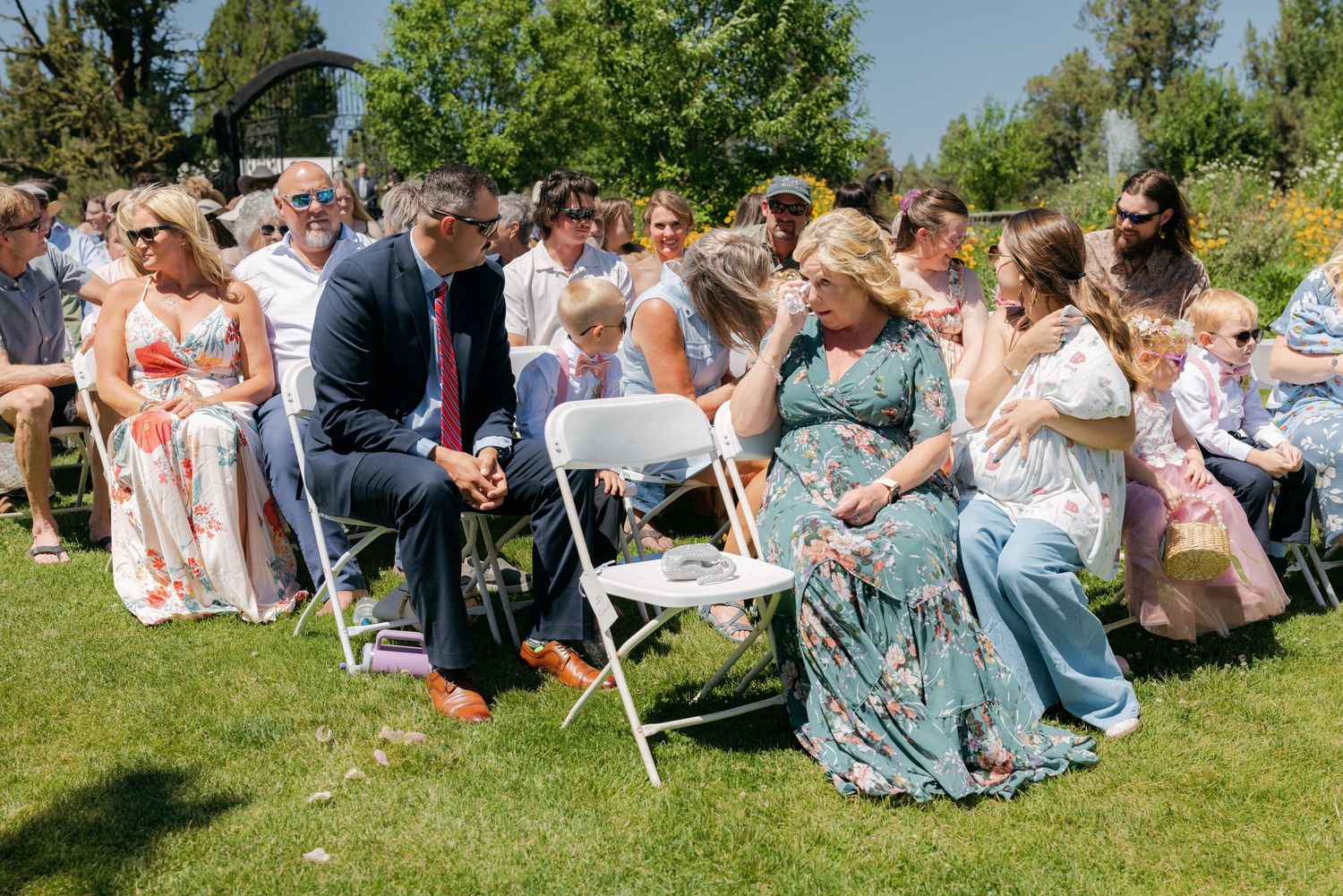 Wedding guests seated outdoors on white folding chairs enjoying a sunny garden ceremony.