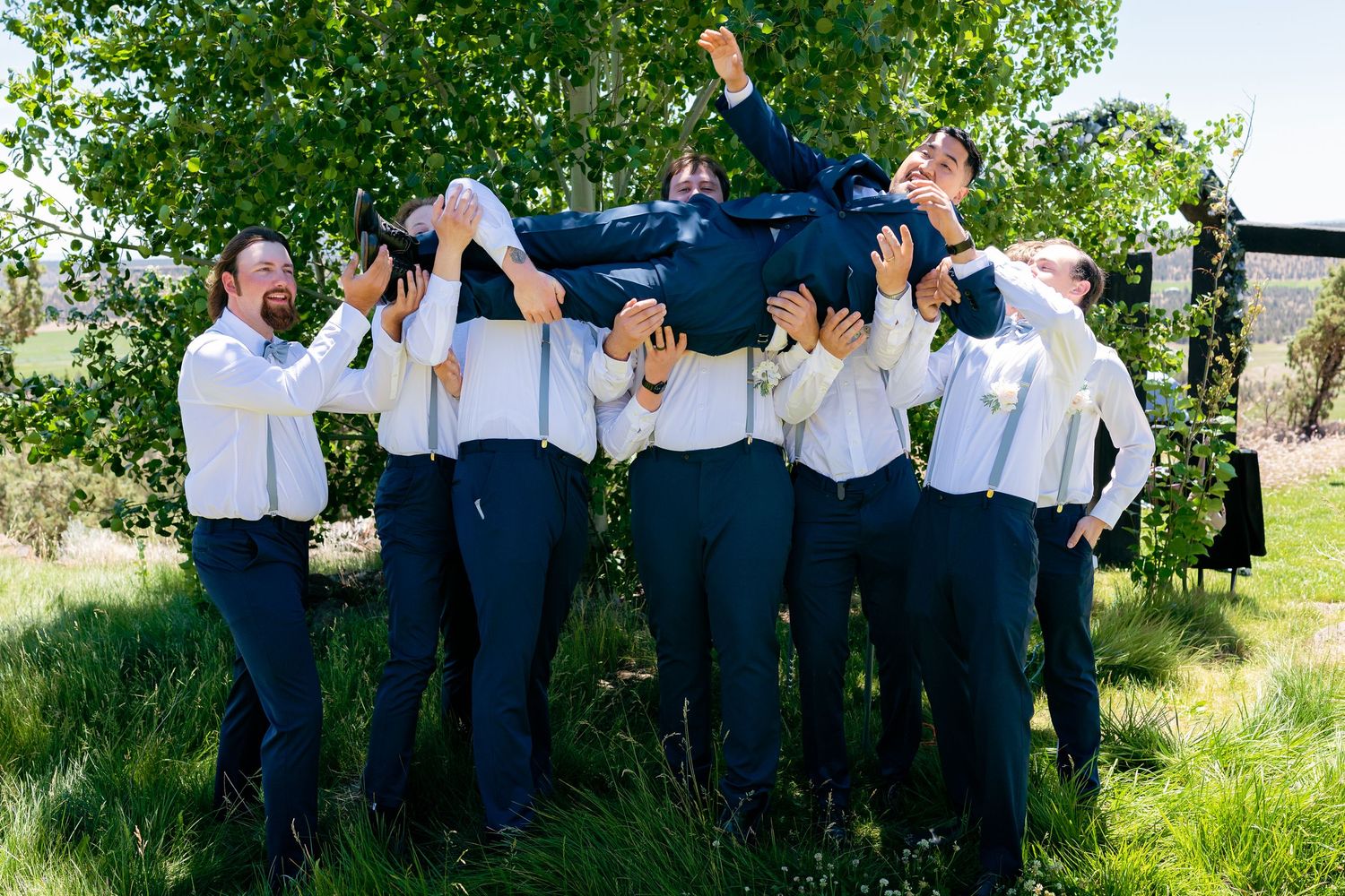 Groomsmen in white shirts lift groom overhead while celebrating outdoors at a wedding.