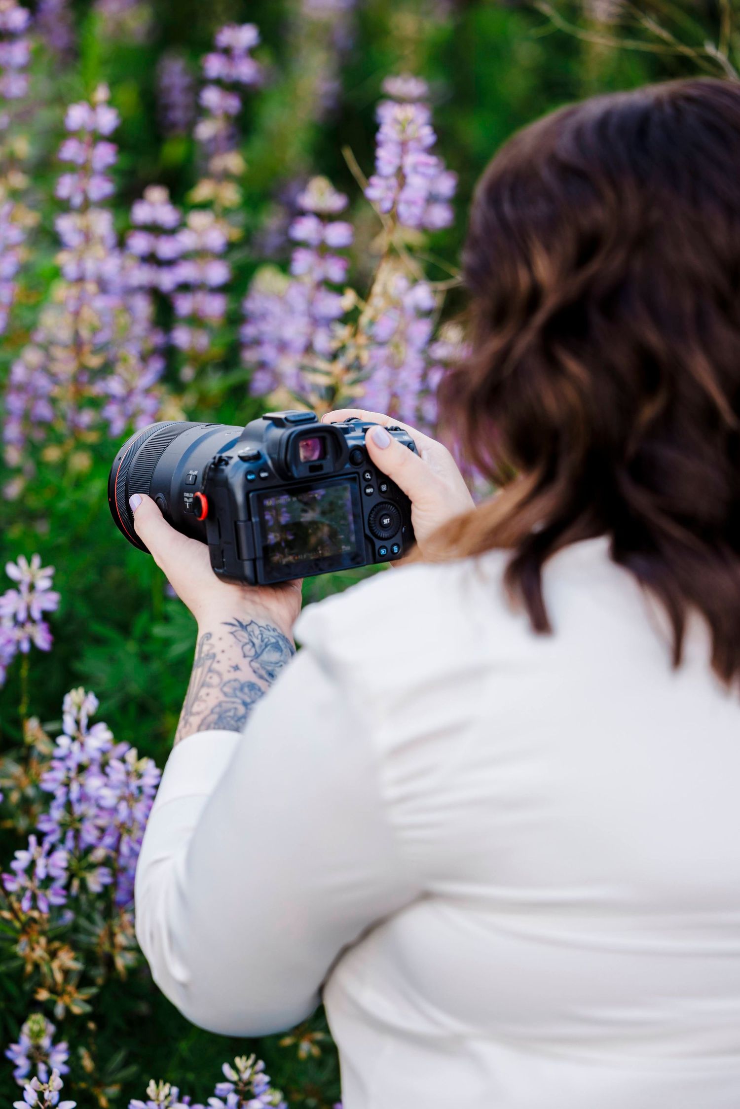 Photographer with tattoo sleeve captures purple lupine flowers using DSLR camera in garden setting.