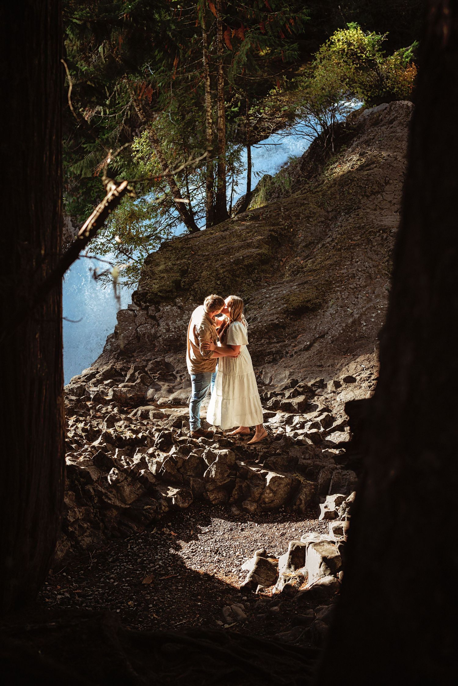 A figure in a flowing white dress stands on a rocky hillside path beneath towering pine trees in dramatic lighting.