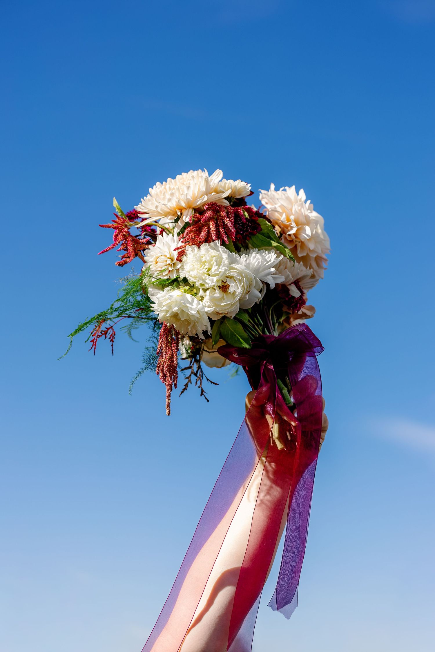 A bridal bouquet with white flowers and red accents held up against a bright blue sky with flowing ribbons.