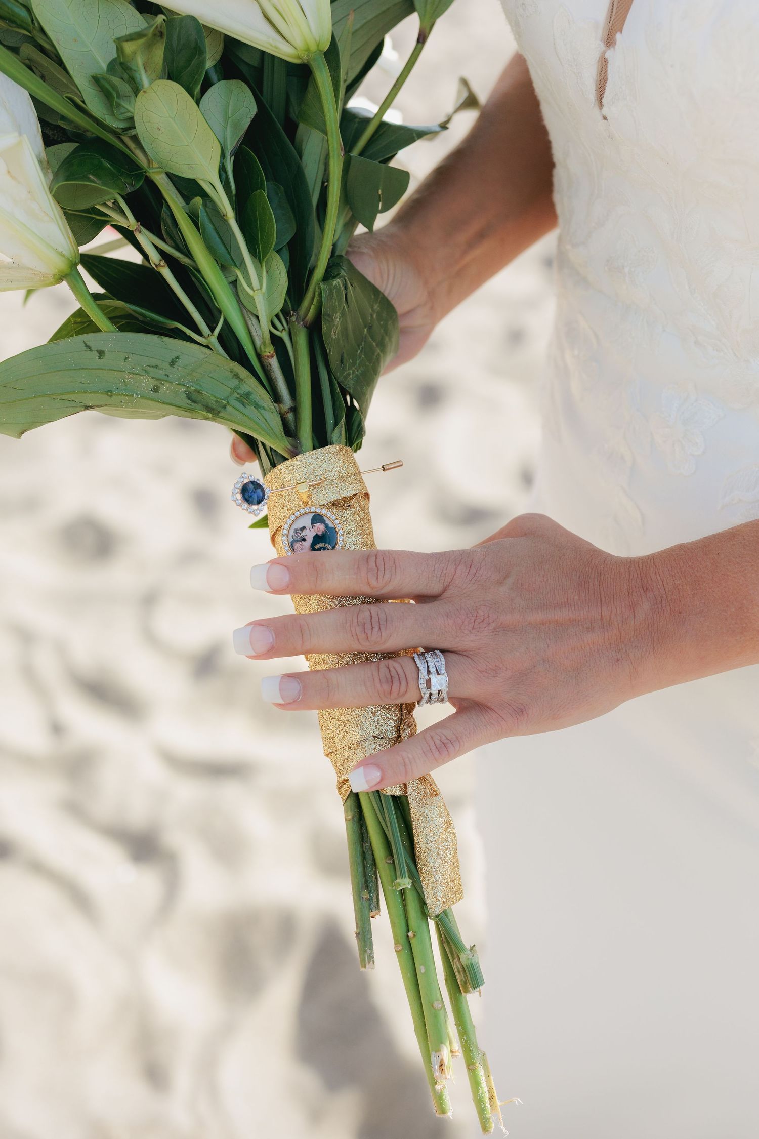 Close-up of hands holding white floral bouquet with wedding rings and lace details.