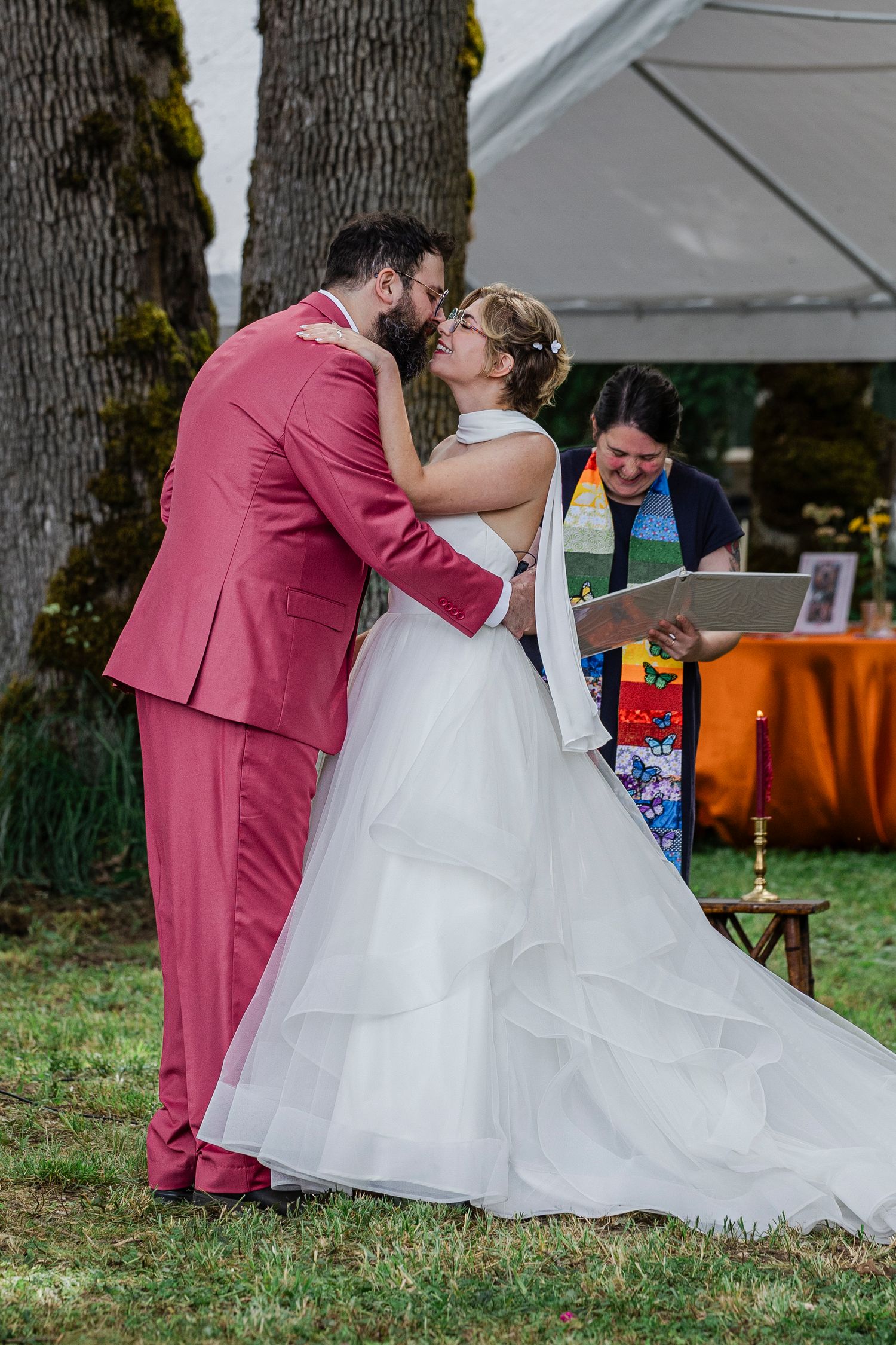 A couple in wedding attire share a romantic moment outdoors in front of trees while an officiant looks on.