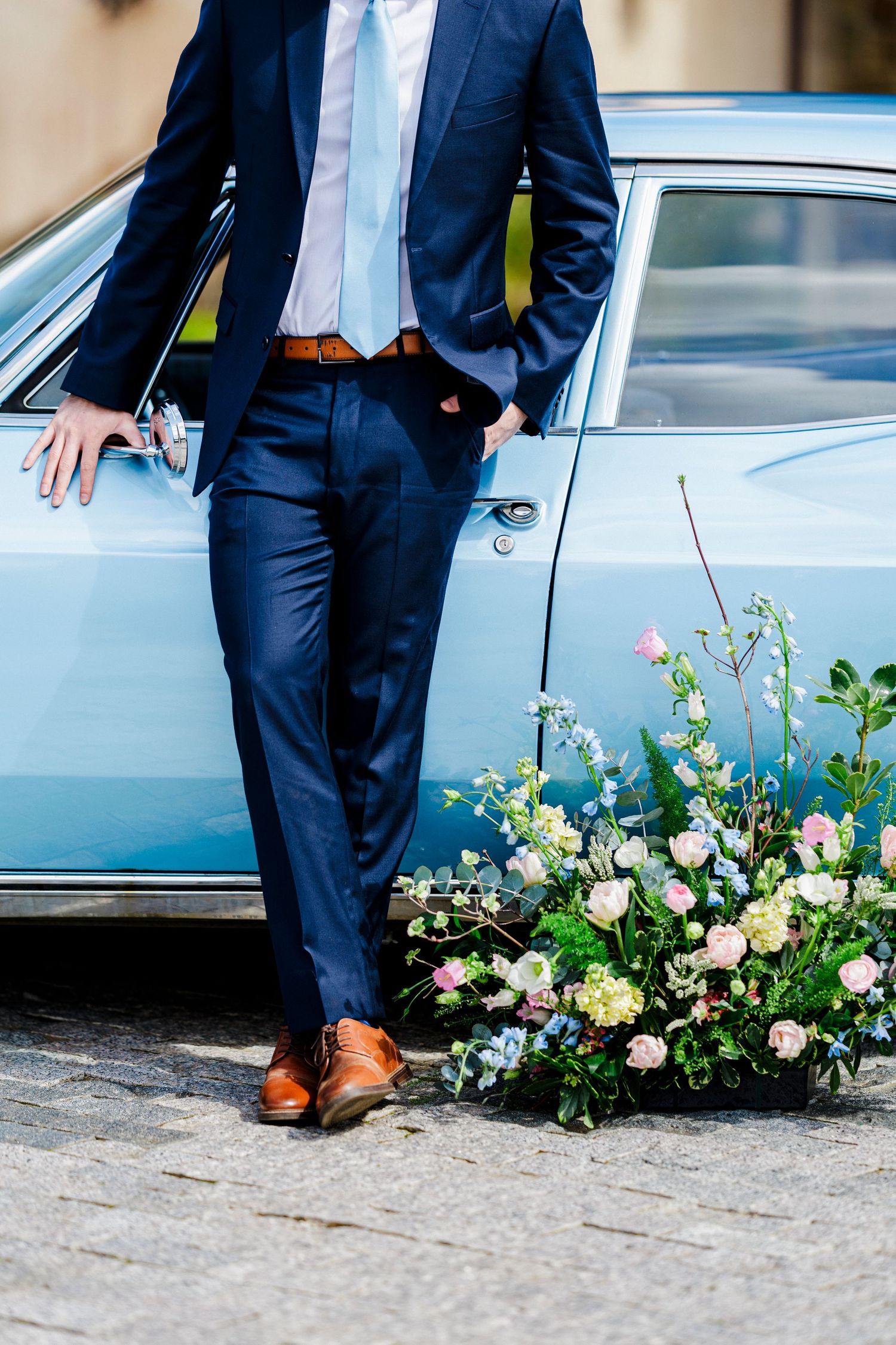 Navy blue suited figure stands beside vintage car with brown leather shoes and floral arrangement at base.