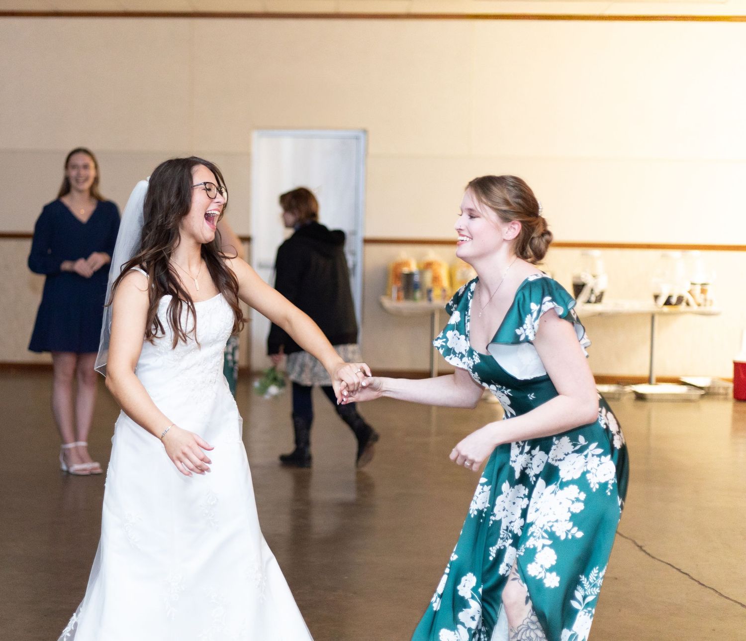 Two people dance together joyfully at a celebration, one wearing a white wedding dress and another in a green floral dress.