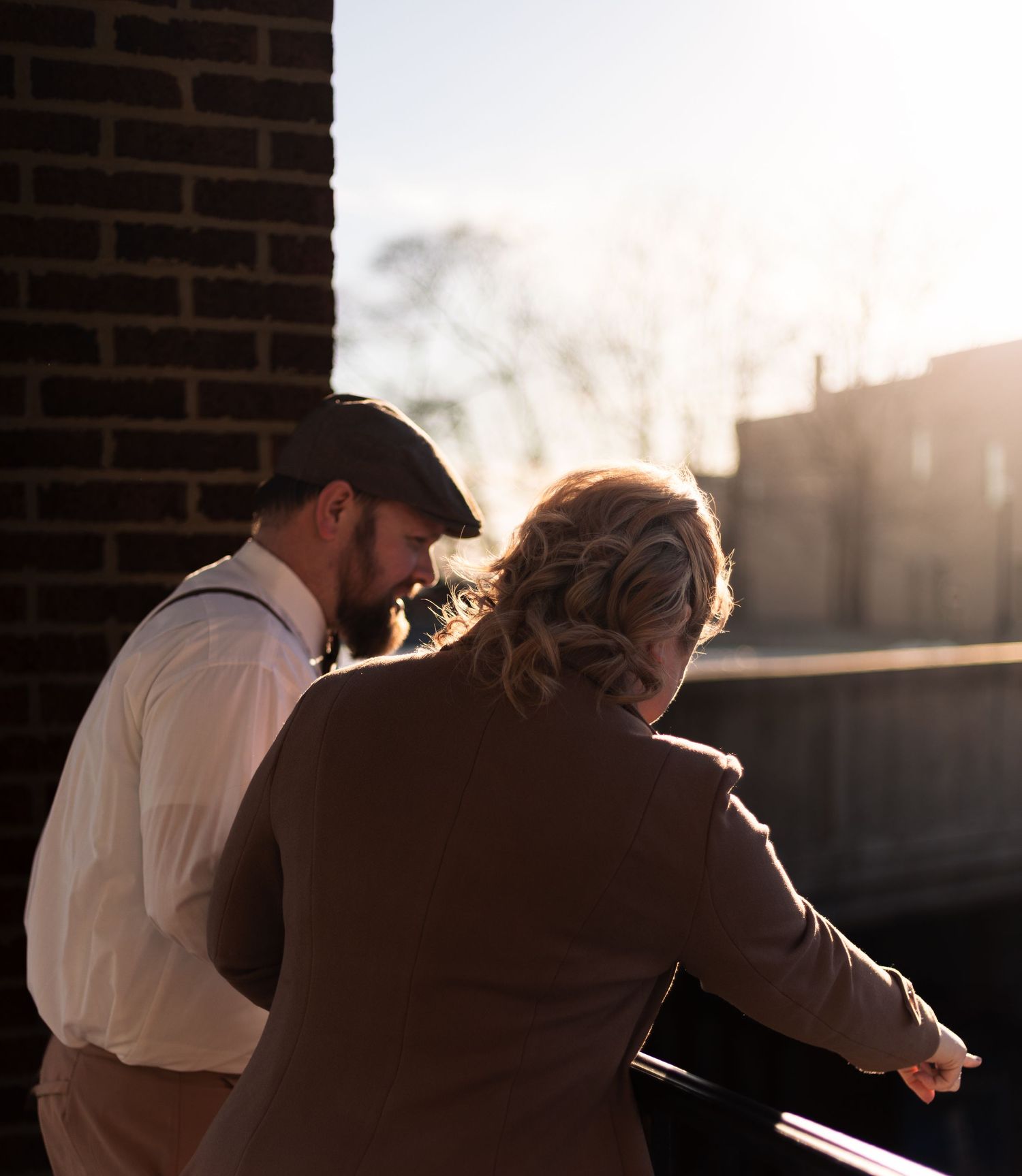 A man in suspenders and flat cap leans toward a woman in golden backlit sunset near a brick wall.