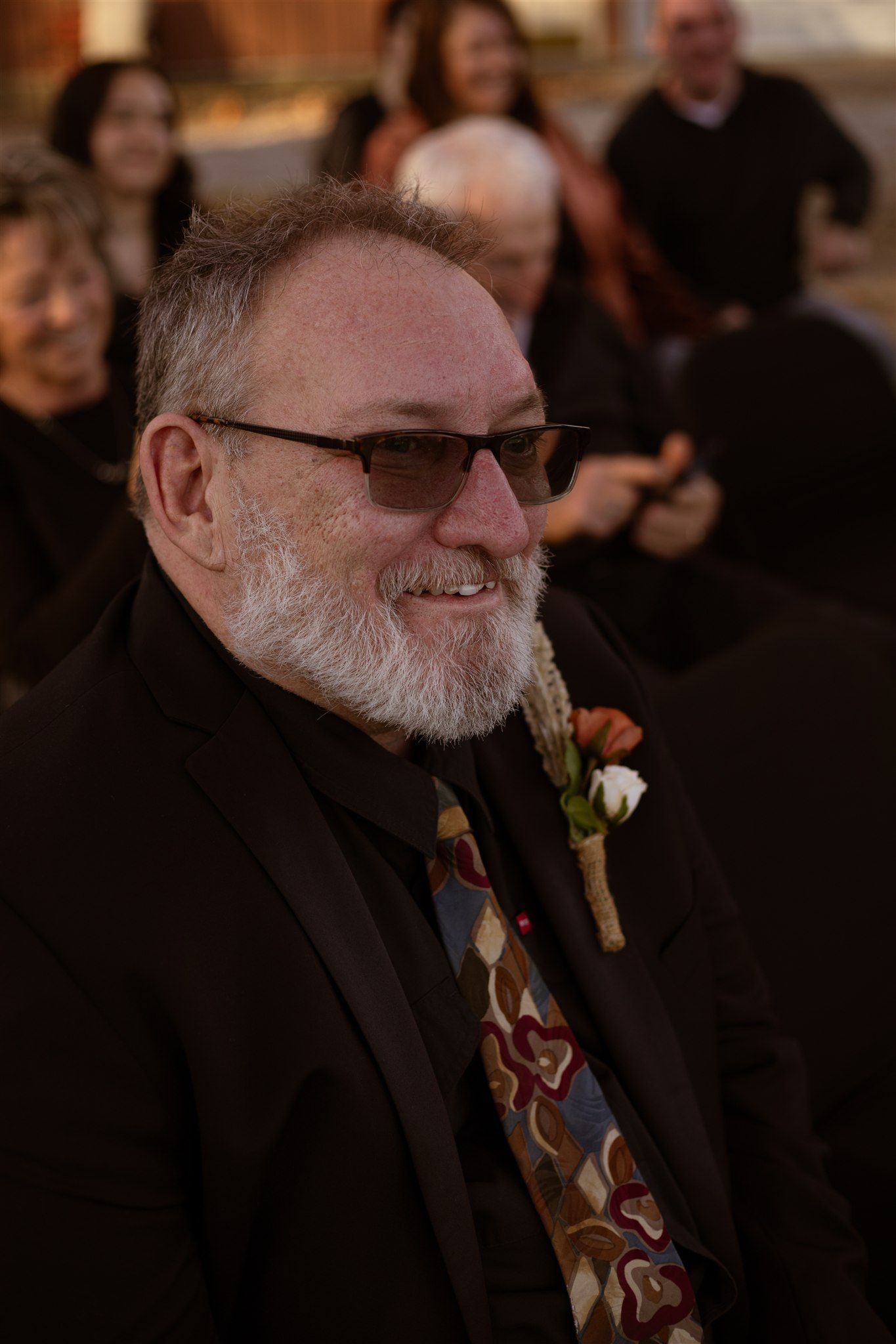 Wedding guest in black suit and tie smiling warmly during outdoor celebration.