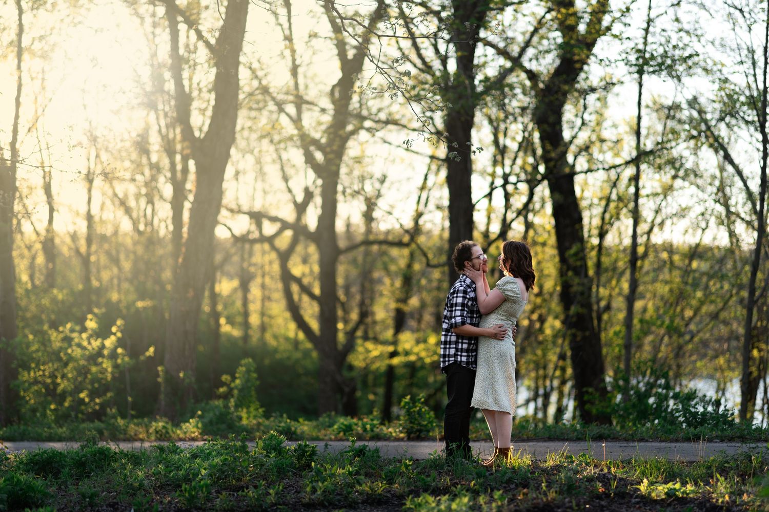 Couple embracing in a sunlit park with golden light filtering through bare trees and lush green shrubs in spring.