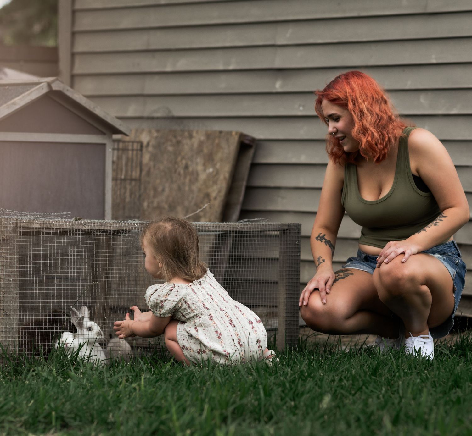 A red-haired woman crouches beside a toddler girl petting a rabbit in a backyard cage on green grass.