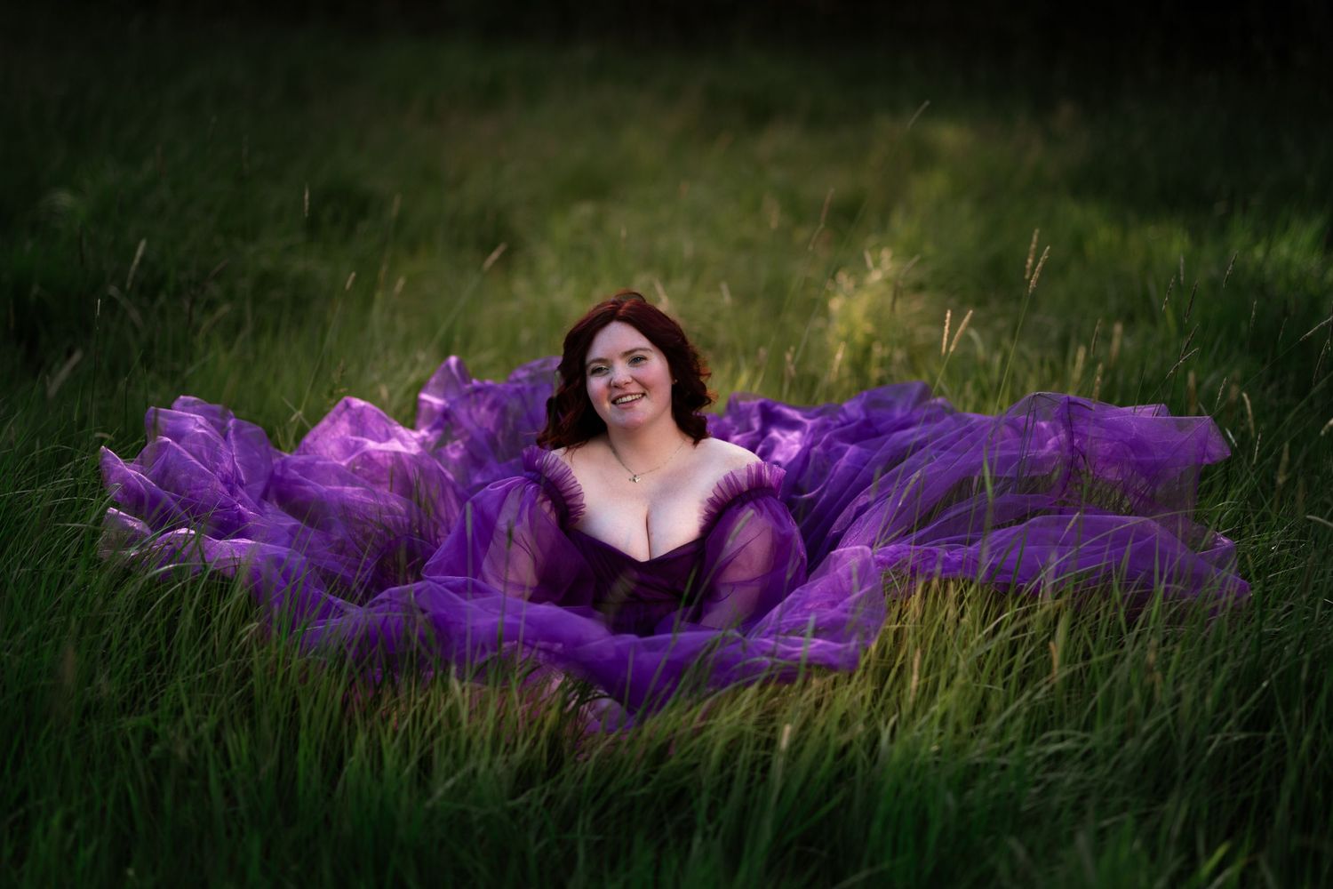 Woman in flowing purple gown sitting in lush green field, smiling gracefully during outdoor portrait session.