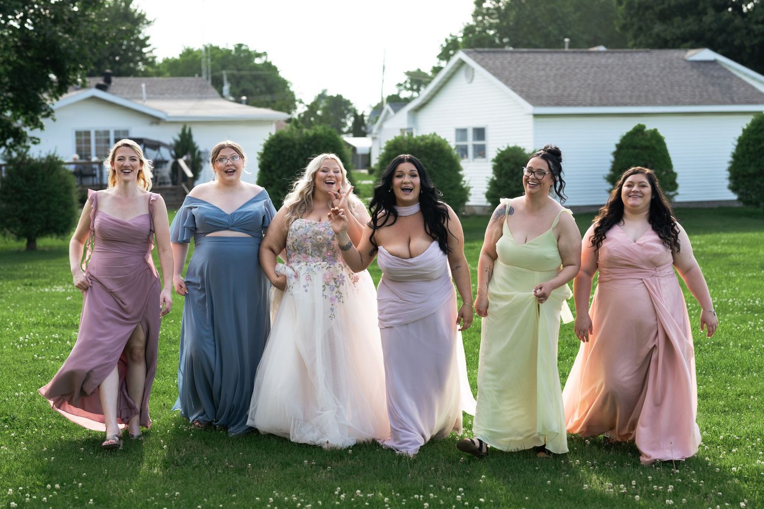 Six women in formal gowns, including a bride in white, smile and walk together on green grass outdoors.