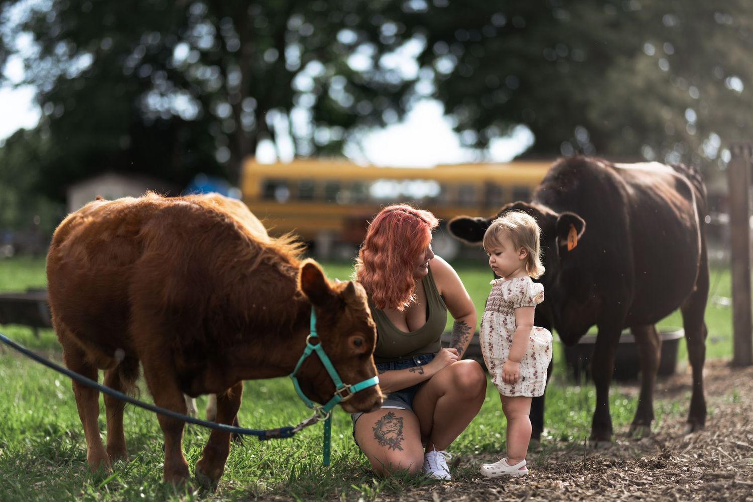 Woman with red hair and tattoos crouches with toddler petting brown calf on green farm, yellow school bus in background.