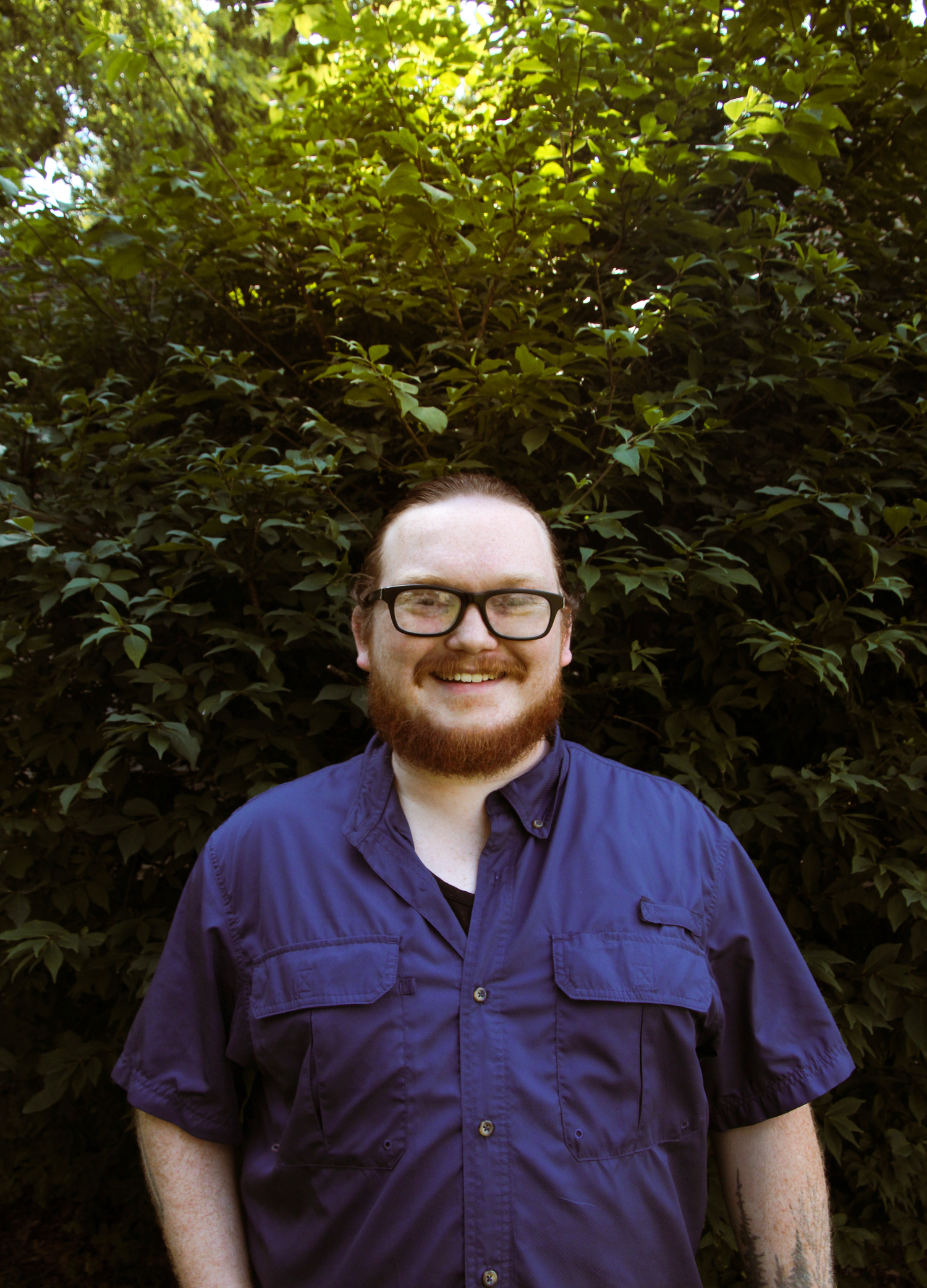 Smiling man with red beard and glasses wearing a blue shirt, standing outdoors in front of lush green foliage.