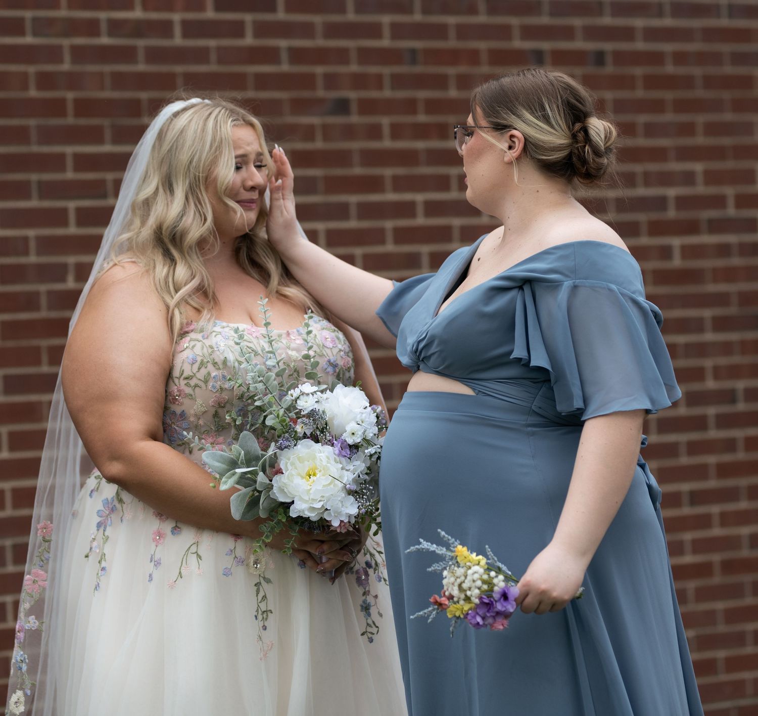 A bride in floral gown with veil holds bouquet as bridesmaid in blue dress gently touches her face near brick wall.