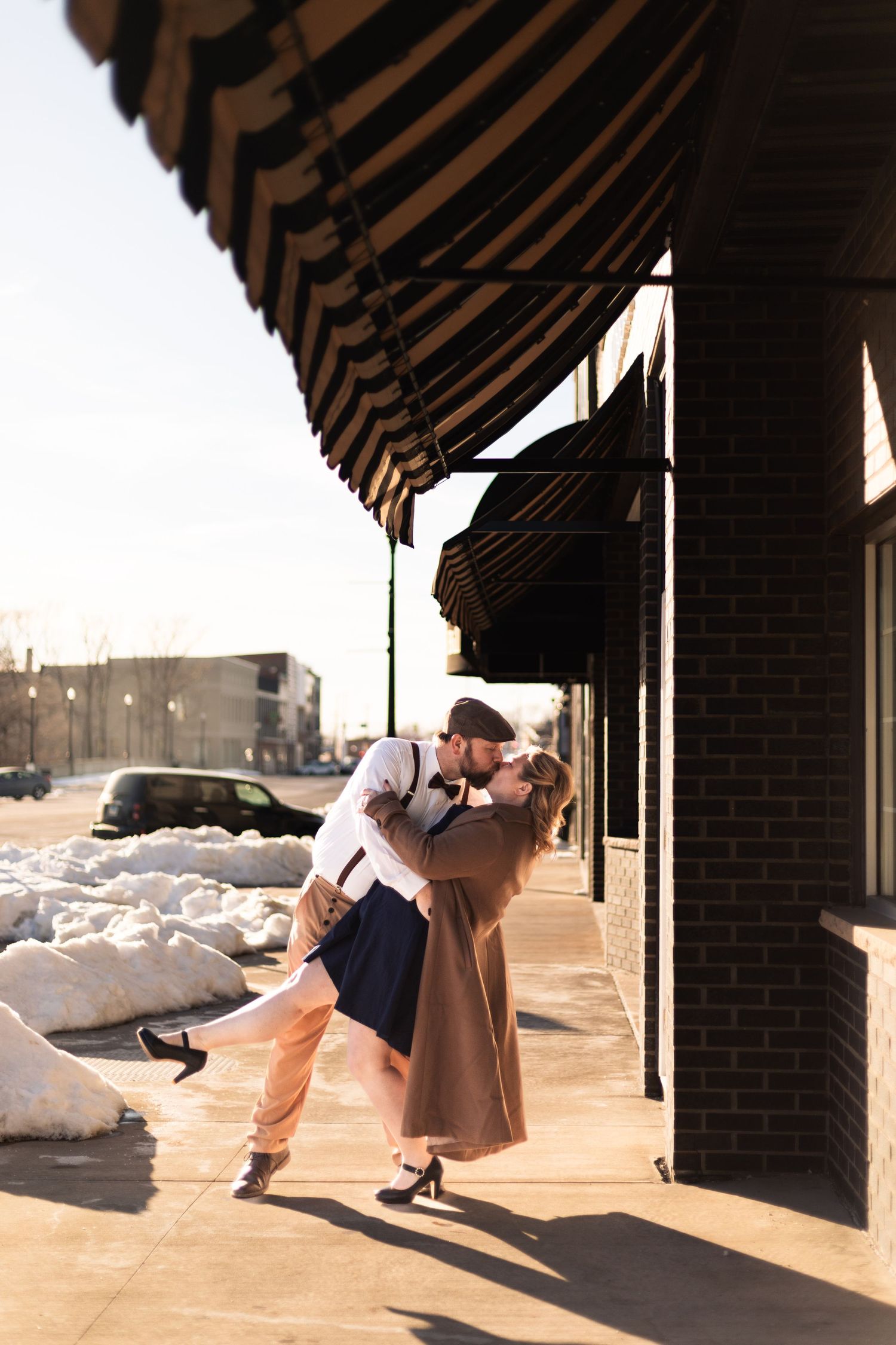 Couple sharing a romantic dip kiss on a sunny sidewalk under striped awnings, with snow piles visible in background.
