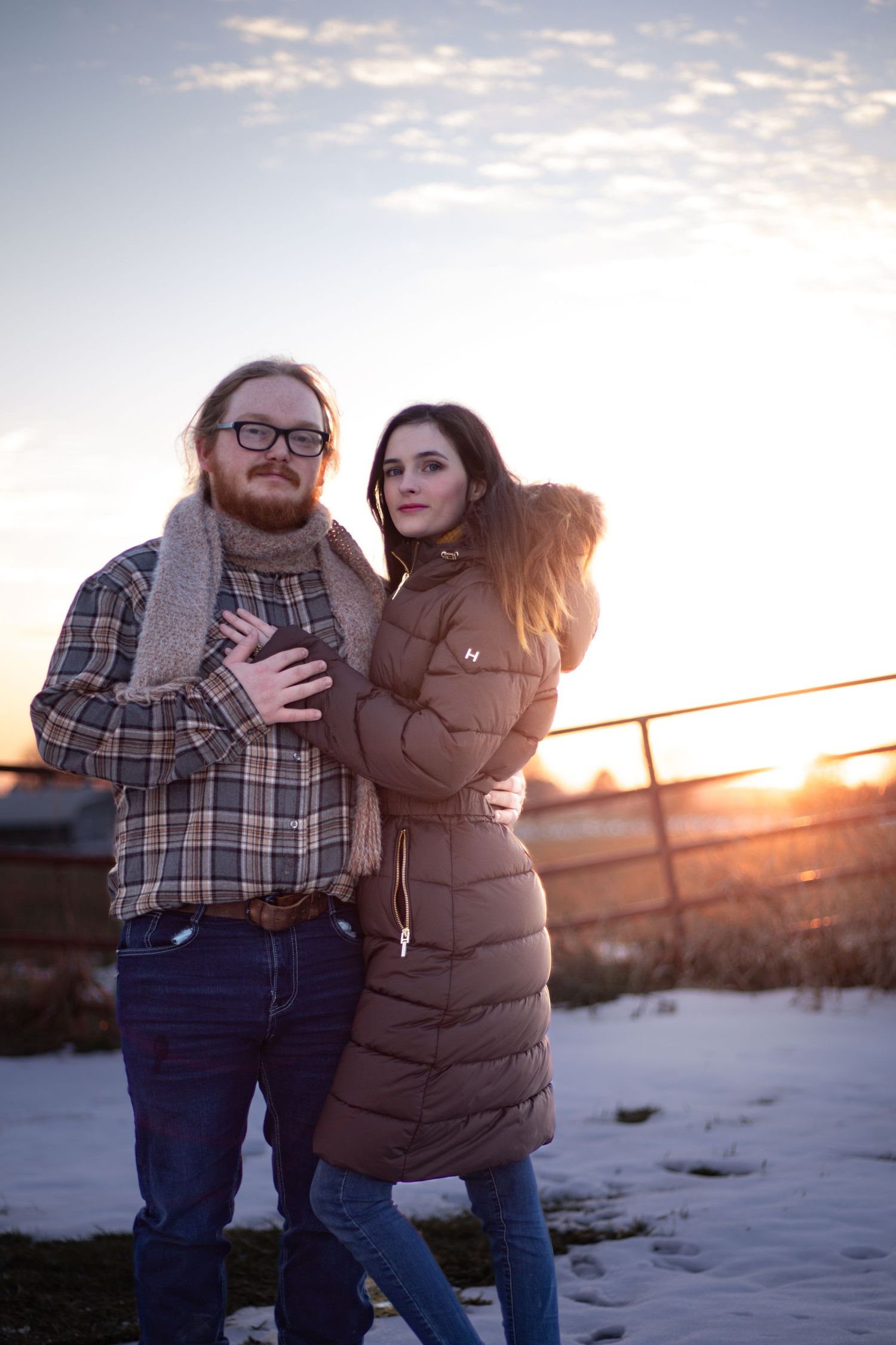 Couple embracing outdoors in winter at sunset, man in plaid shirt and scarf, woman in brown puffer coat, snowy field background.
