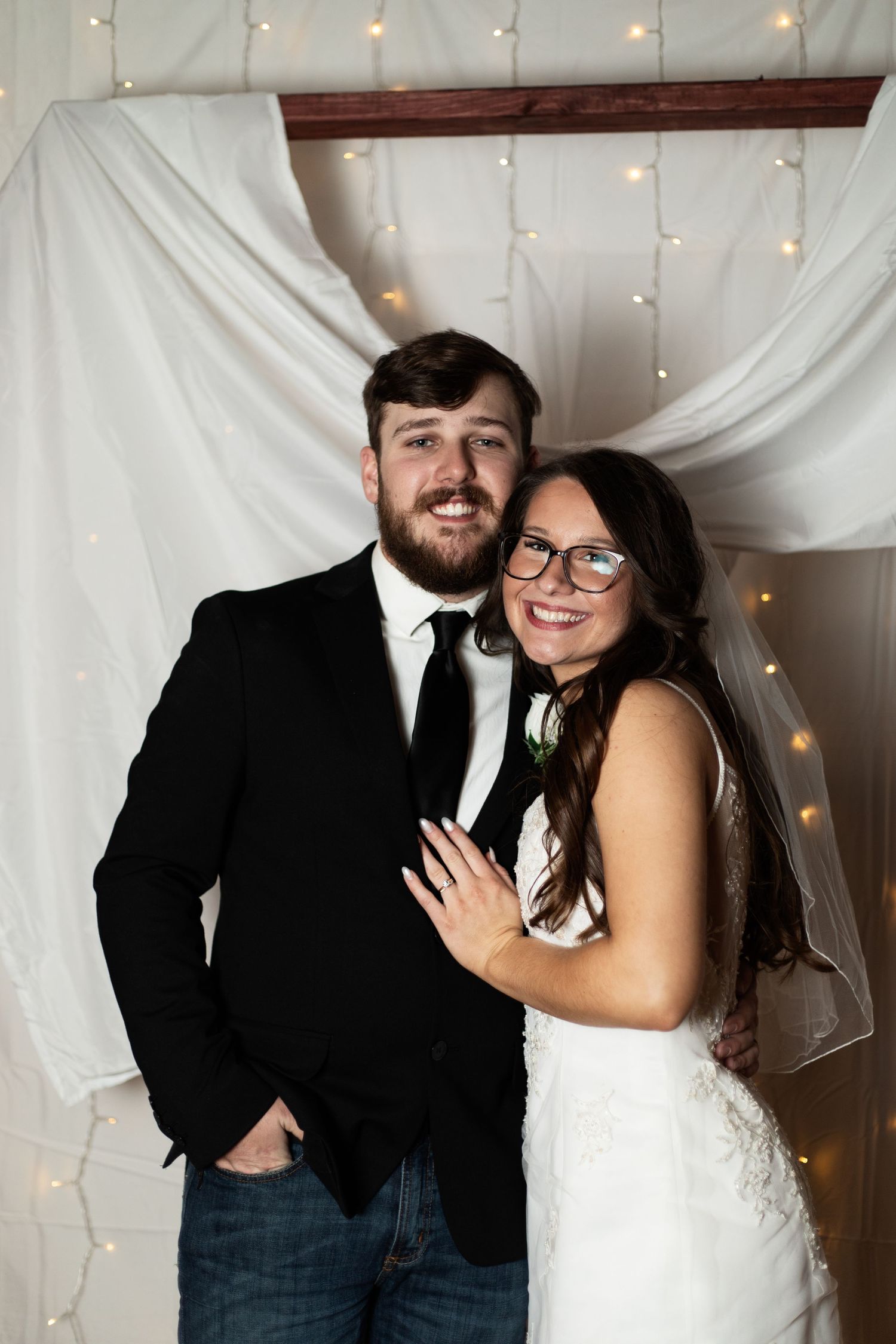 A happy couple poses together against a white backdrop with twinkling string lights, the bride wearing a lace gown and veil.