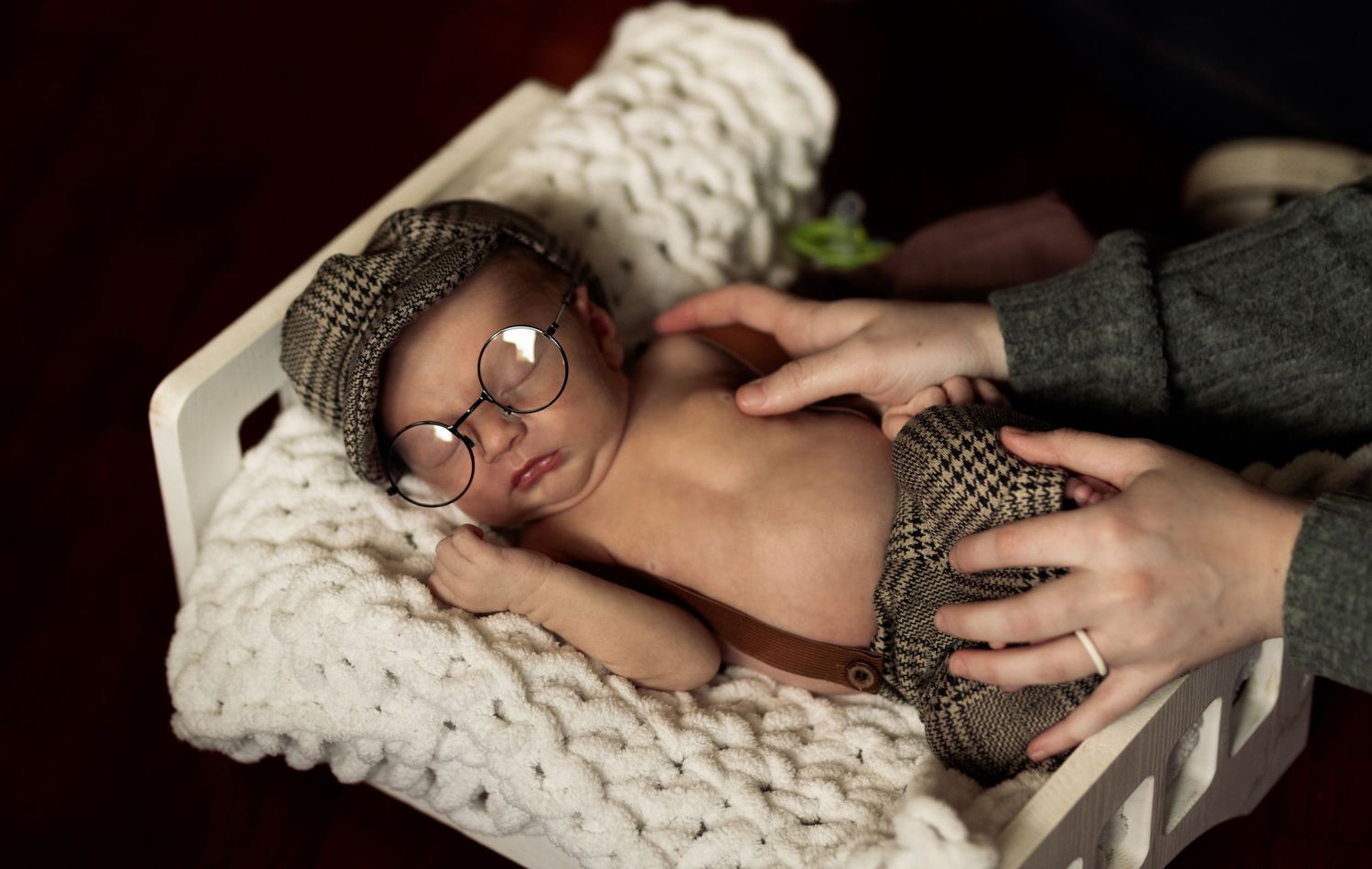 Newborn baby wearing tiny glasses and plaid hat sleeping in white wooden crate on knit blanket.