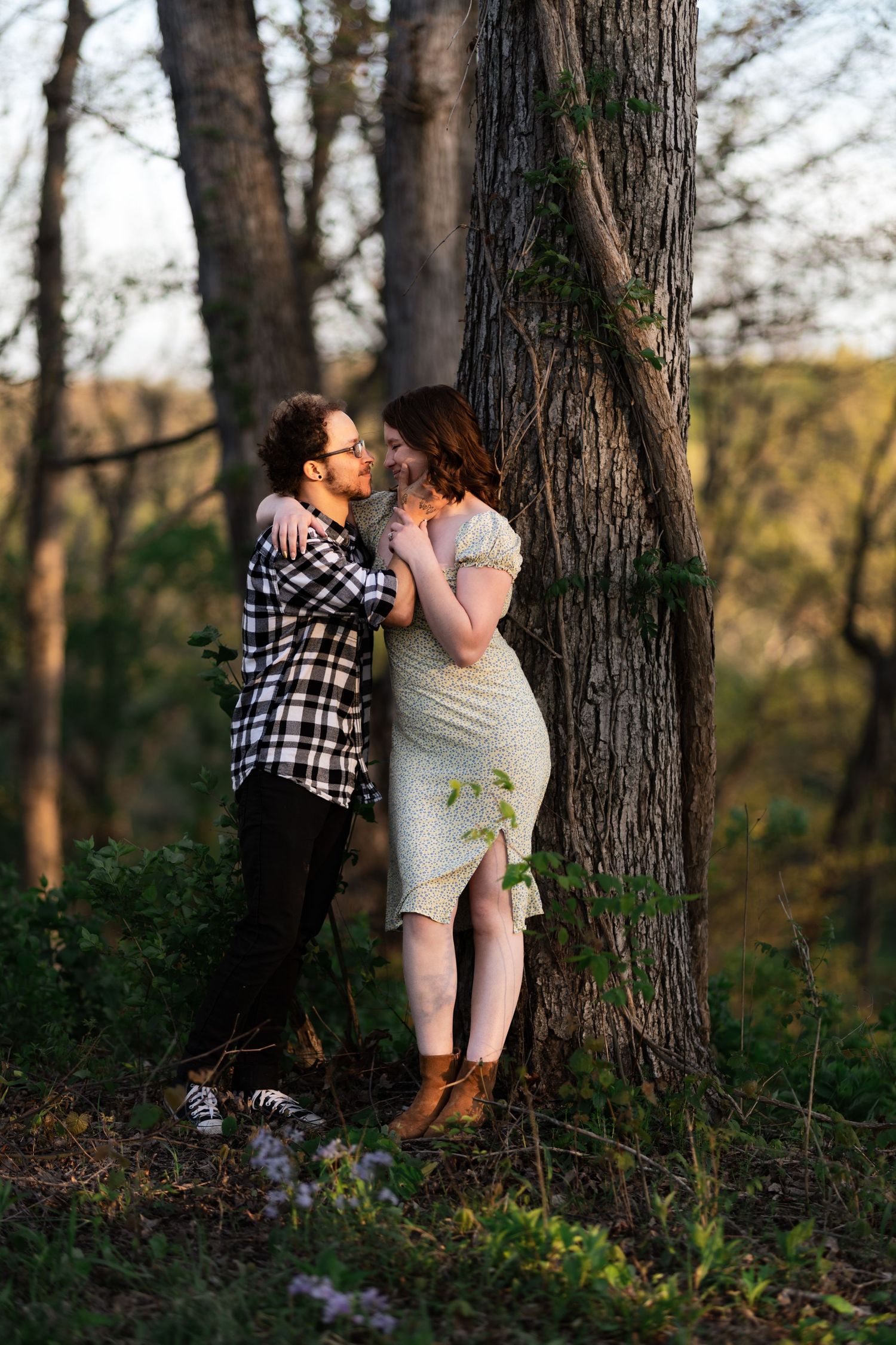 Couple sharing a romantic moment leaning against a tree in a sunlit forest, woman in white dress and man in plaid shirt.