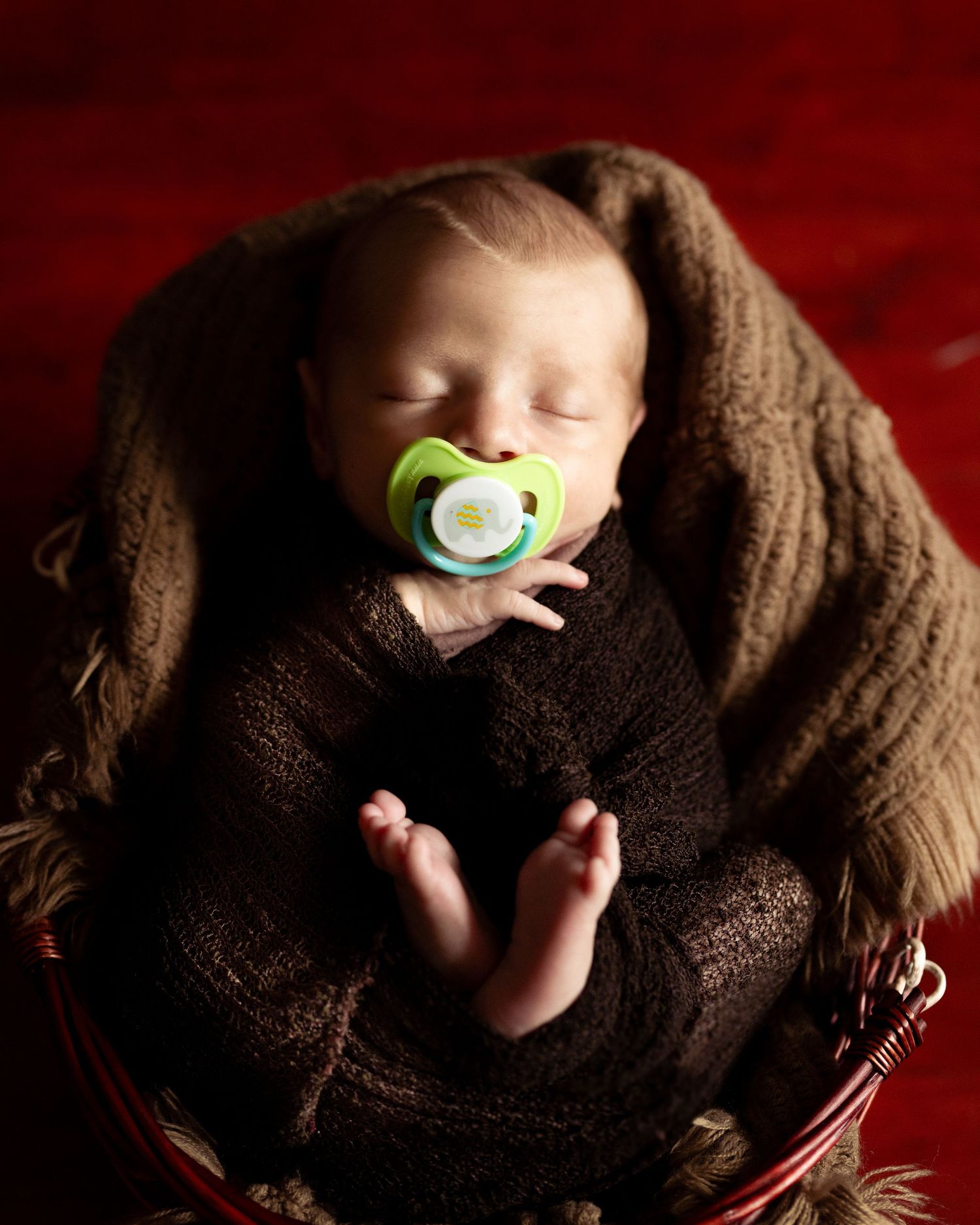 Sleeping newborn baby wrapped in brown knit blanket with green pacifier, nestled in basket on red background.