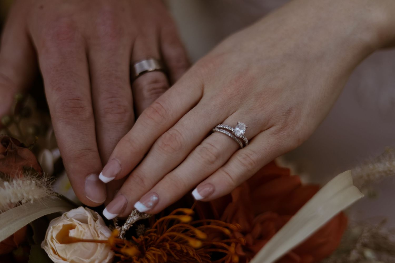 Couple displaying wedding rings on hands resting over autumn floral bouquet with roses in warm tones.