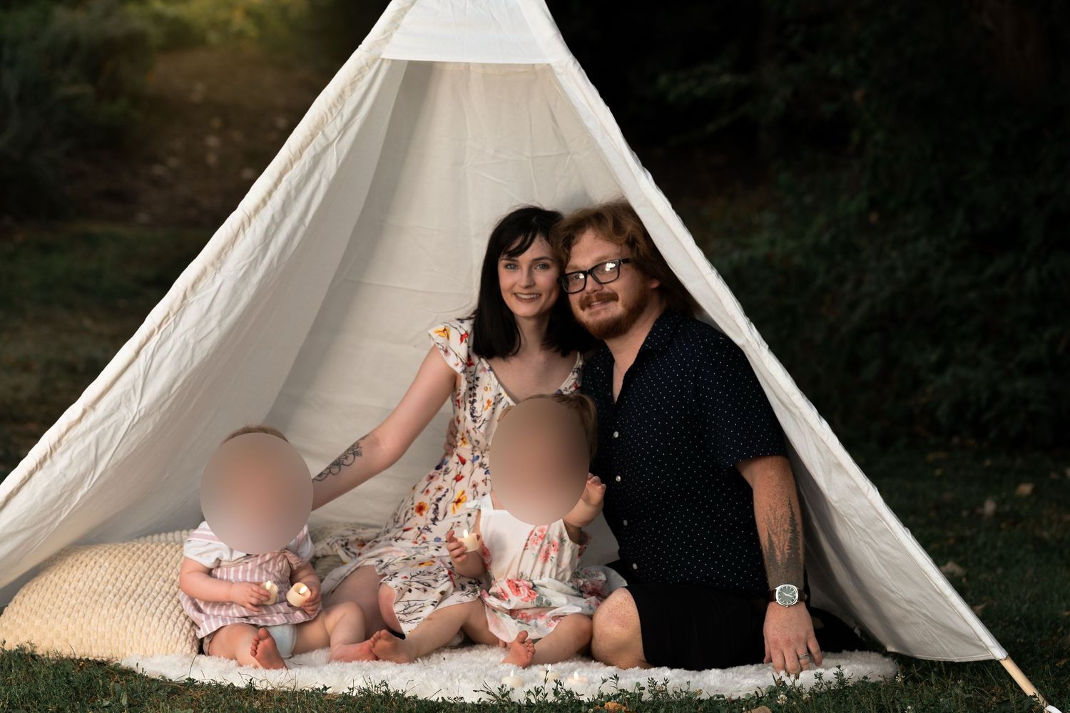 Family of two adults and two young children sitting together under a white teepee tent outdoors.