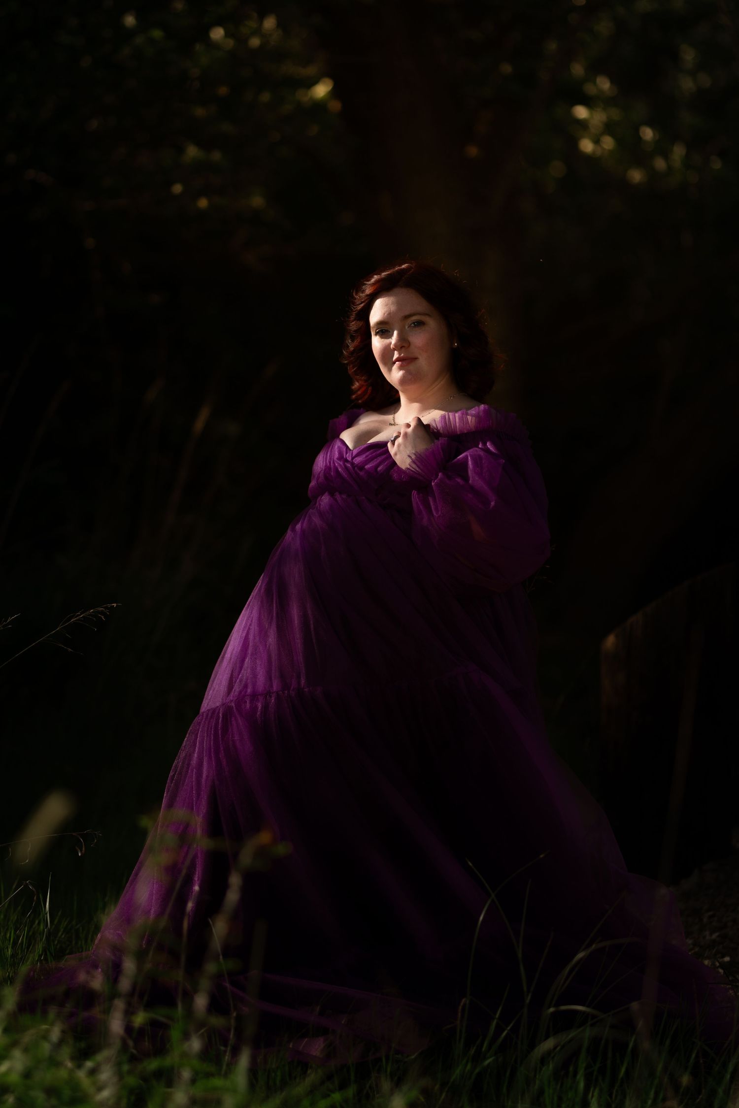 Woman in flowing purple tulle gown standing in a dark forest, bathed in soft golden light.