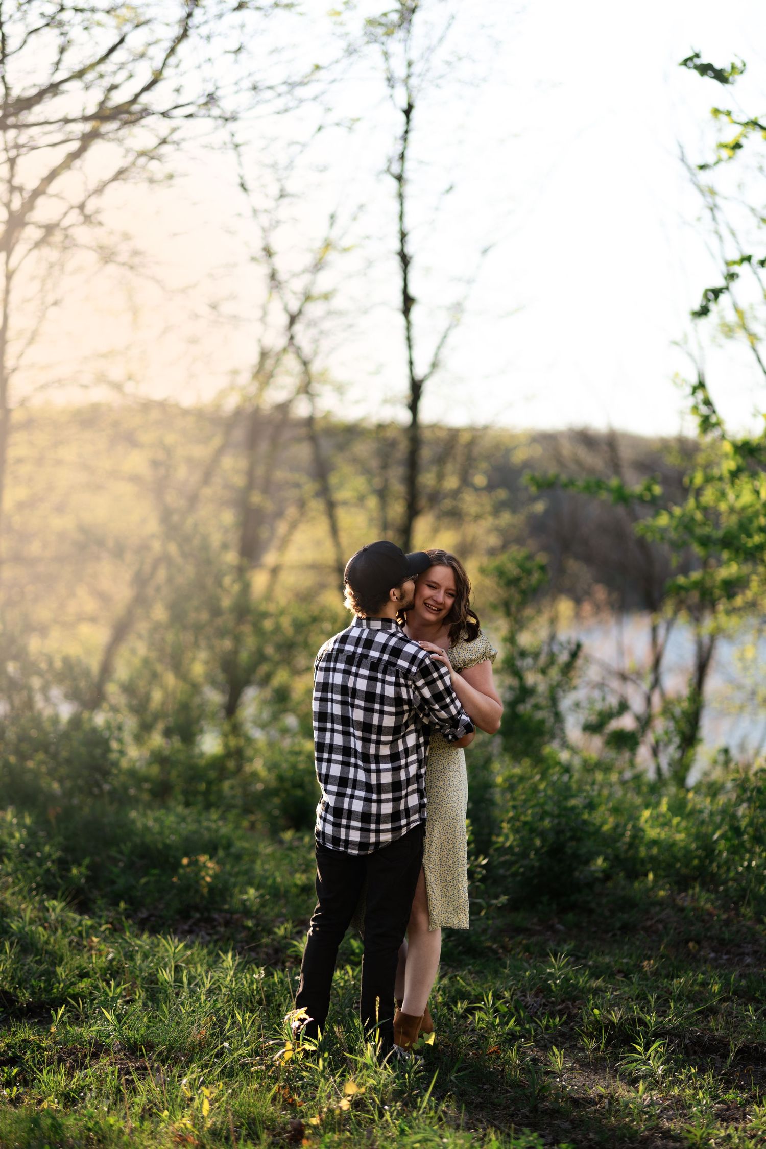 Couple embracing outdoors near a sunlit lake, surrounded by lush green trees during golden hour.
