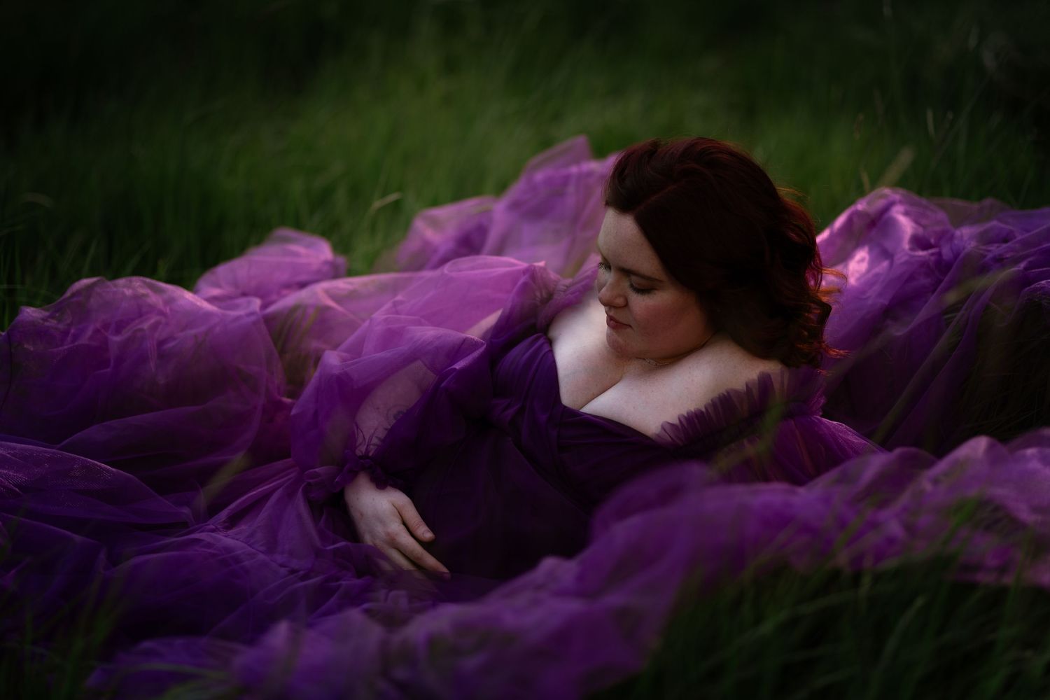 Pregnant woman in flowing purple gown lying in green grass for maternity photo session.
