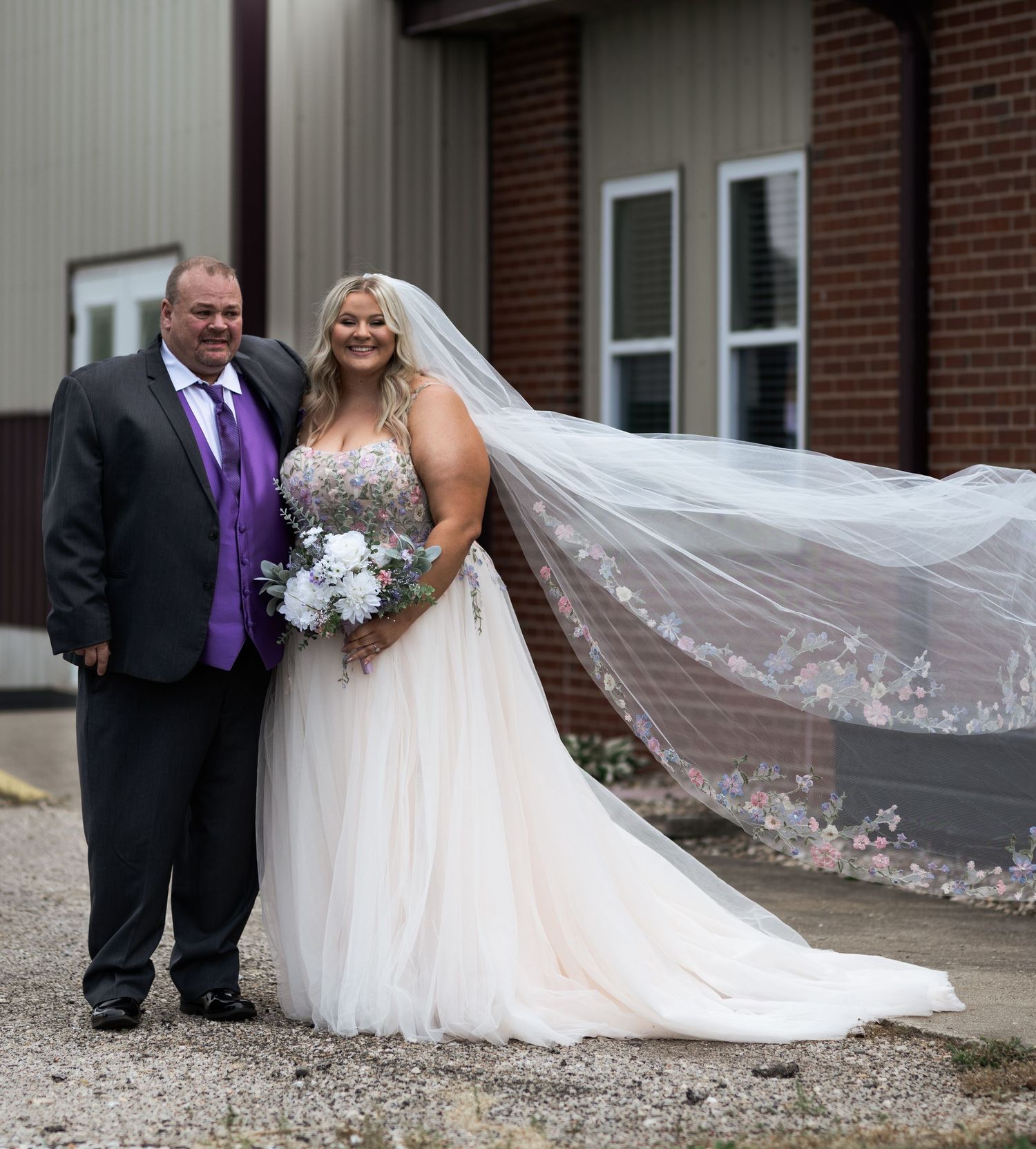Bride in flowing white gown with veil and groom in black suit with purple tie pose outdoors on wedding day.