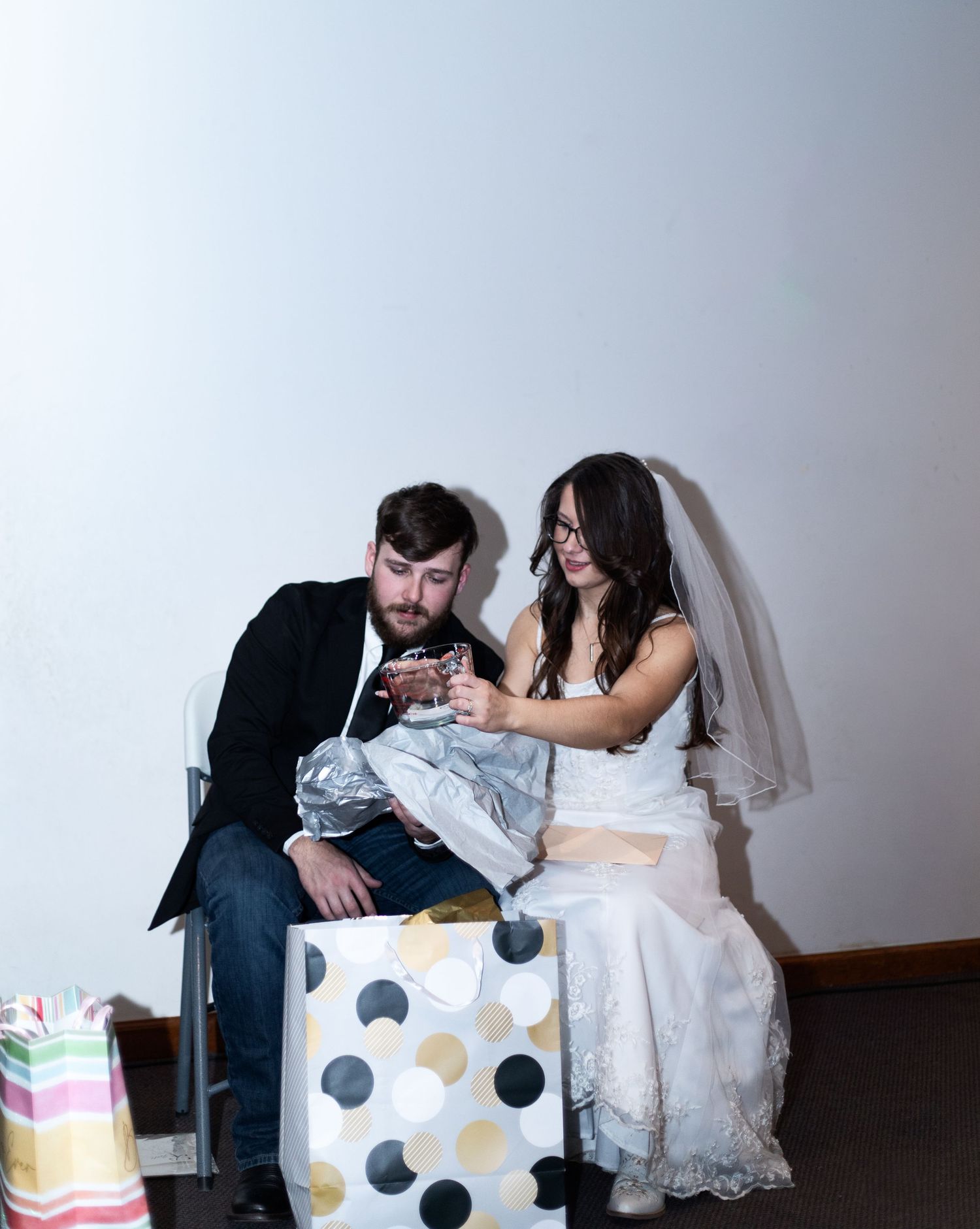 A bride in a white gown sits beside wedding gifts during a reception celebration.