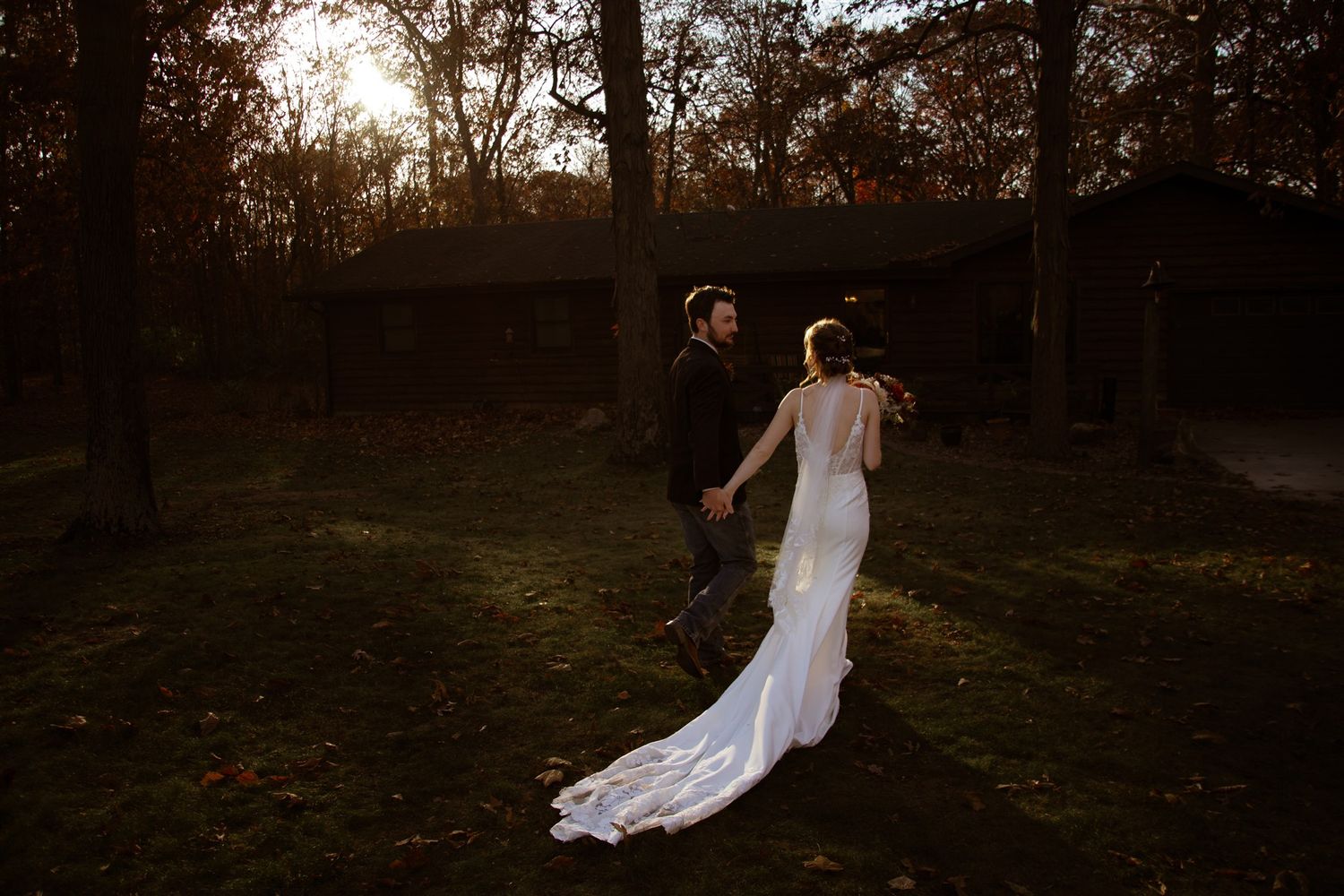 A couple in wedding attire runs playfully through an autumn forest at sunset.