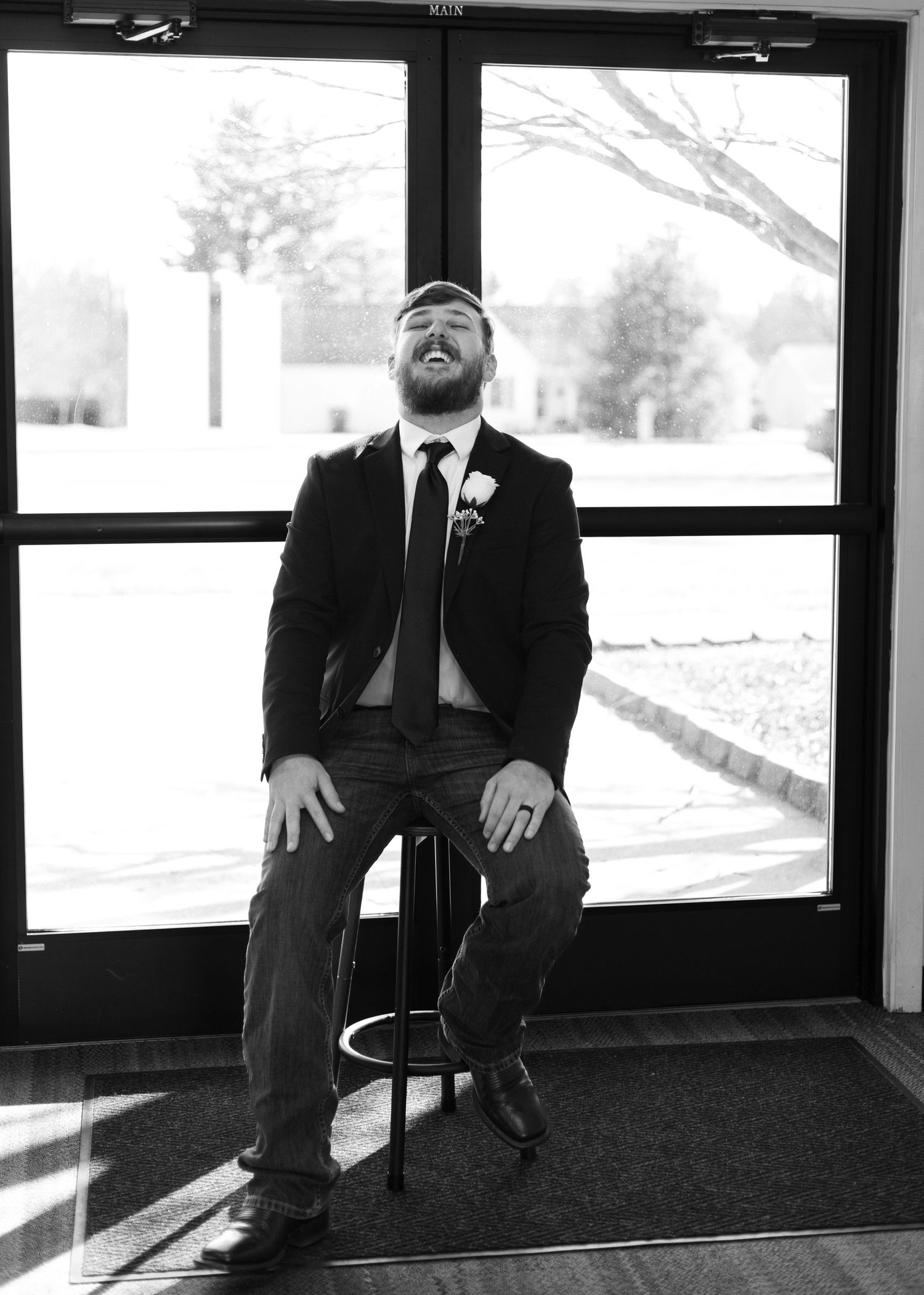 Man in formal attire sits alone in waiting area with large windows overlooking snowy winter scene.
