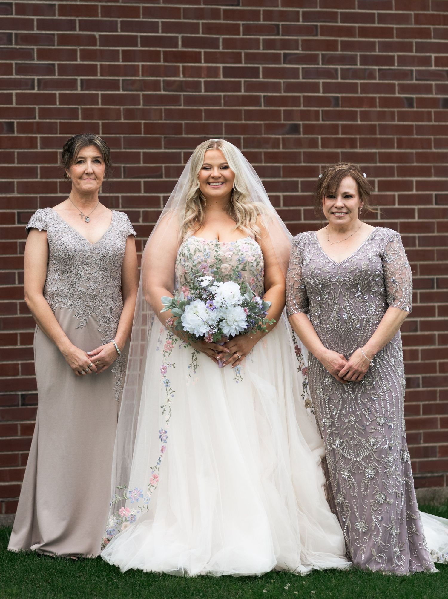 Bride in white gown holding bouquet stands with two bridesmaids in silver beaded dresses before brick wall.