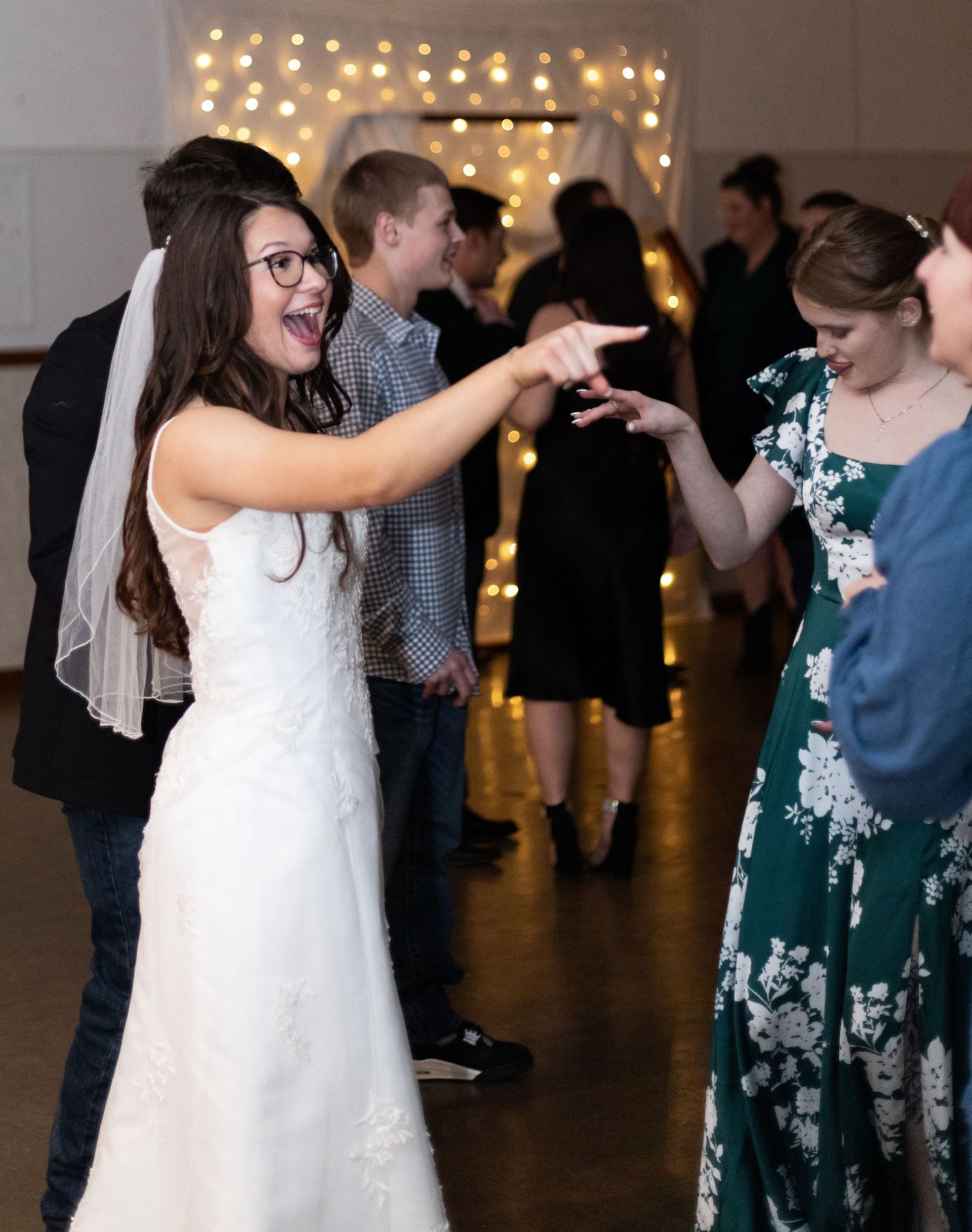 Wedding guests dancing together at a reception with string lights glowing overhead in an indoor venue.